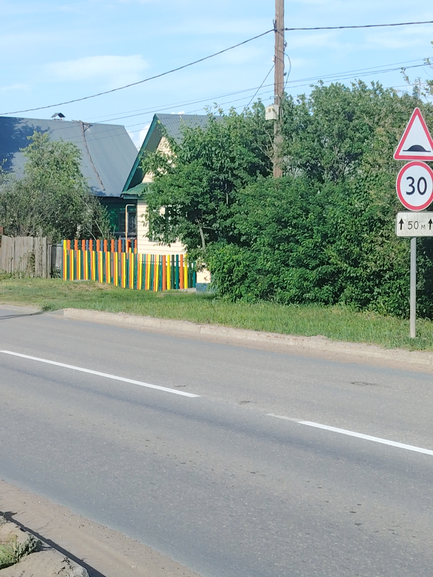 The image shows a street scene in what appears to be a residential area. A paved road with a white lane marking runs horizontally across the lower portion of the frame. A sidewalk or grassy verge borders the road on the right. Beyond this verge, there is a colorful wooden fence painted with vertical stripes of yellow, green, and orange.
Behind the fence stands a house with a light-colored exterior and a dark roof. Another house with a similar dark roof is partially visible to the left. Lush green trees and bushes are scattered throughout the scene, partially obscuring the house and fence.
A utility pole stands near the right edge of the image. A road sign indicating a speed bump ahead and a speed limit of 30 km/h, with a distance marker of 50 meters, is positioned to the right of the pole, partially obscured by foliage. The sky is light blue with some wispy clouds. The scene suggests a bright, sunny day.
Based on the prompt, this location is in the city of Chaikovsky, Russia.