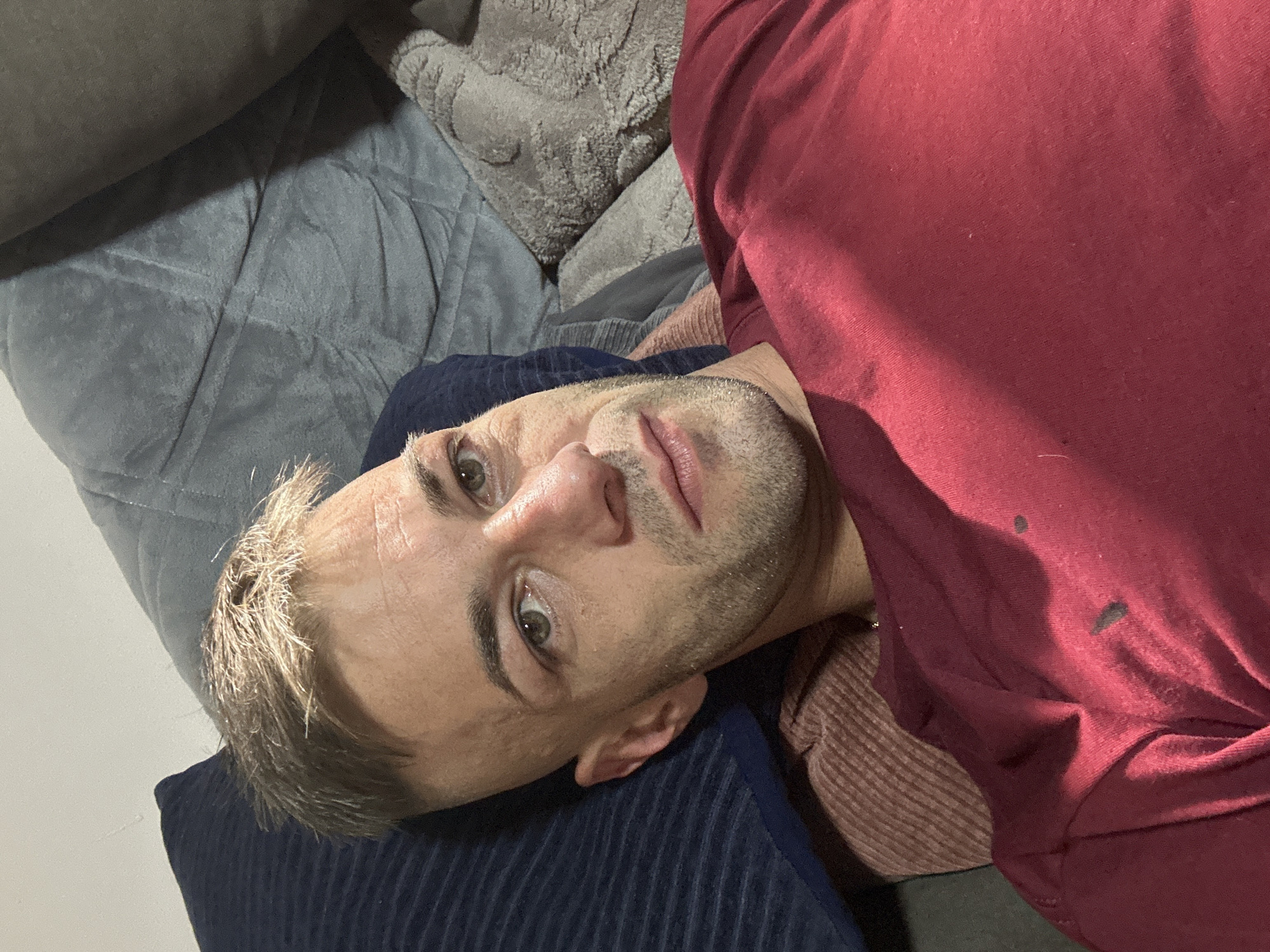 The image shows a close-up, overhead shot of a man lying on a couch. The man has fair skin, light brown hair, and green eyes. He is wearing a maroon t-shirt. The couch appears to be made of a grey, textured fabric, with a dark blue pillow underneath the man's head. The scene is dimly lit, suggesting it is nighttime or indoors with minimal lighting. There is no visible text in the image. The man's expression is neutral. The setting is likely a living room or bedroom in Cherven Breg, Bulgaria.