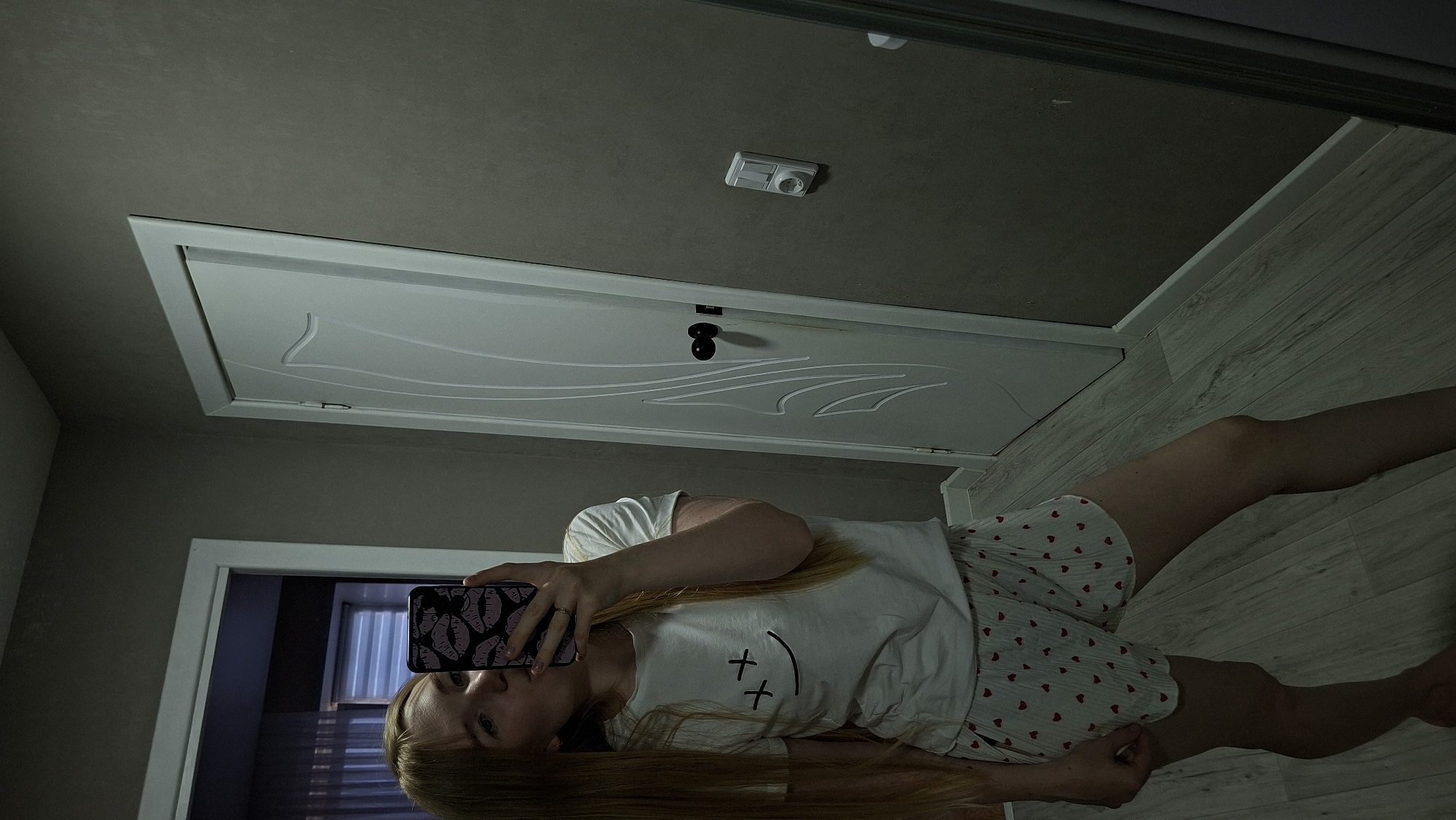 In this full-body mirror selfie, a young woman with long blonde hair is standing in what appears to be a hallway or room with a gray wall and a white door. She is wearing a white t-shirt with a smiley face graphic and white shorts with a heart pattern. The woman is holding a smartphone with a pink and black lip-patterned case, which she is using to take the picture. Her reflection is visible in a large mirror to her right. The flooring is light-colored wood. An electrical outlet and light switch are visible on the wall behind her. The image does not contain information that would allow for its placement in Chaikovsky, Russia.