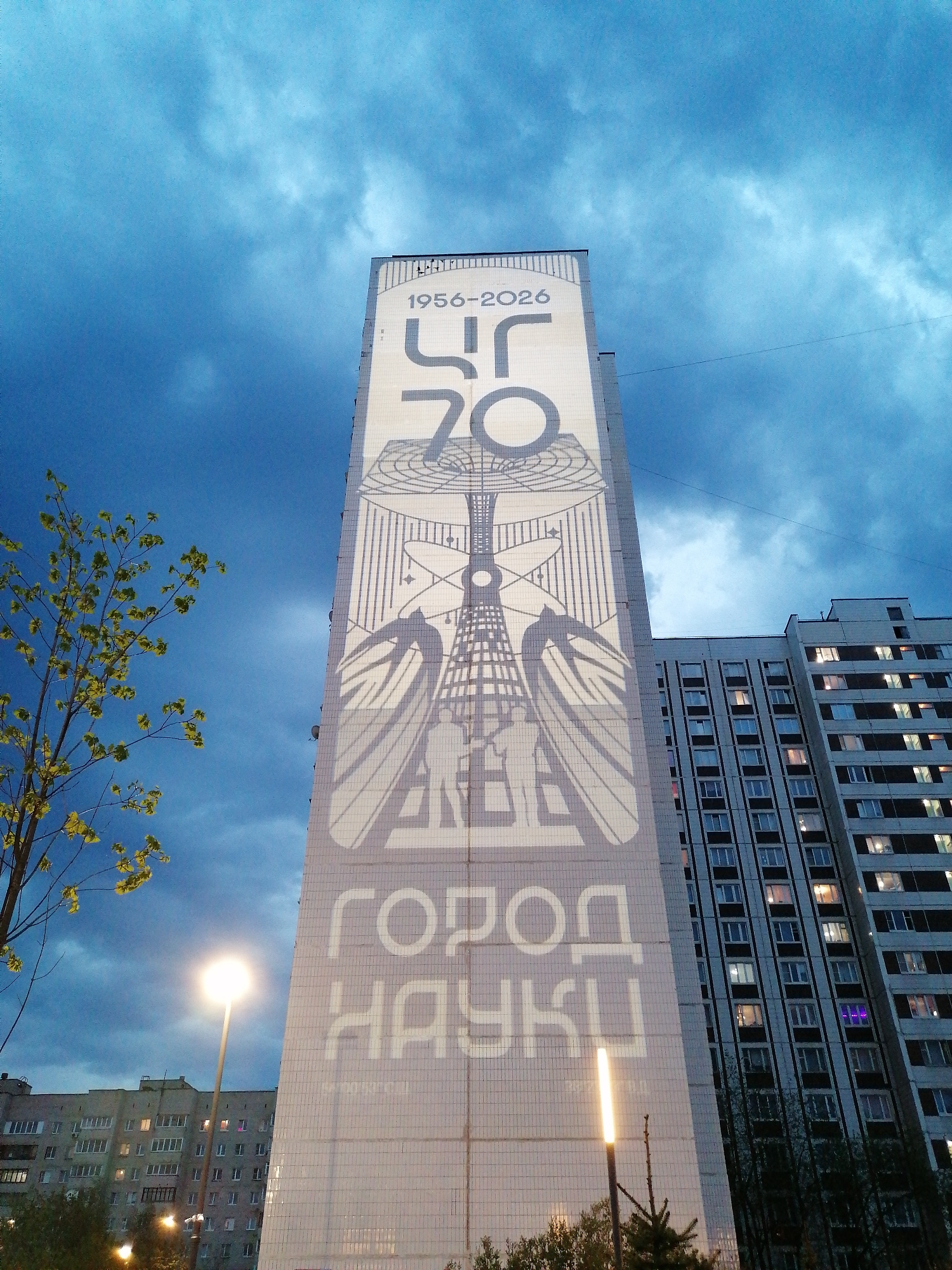 Here's a detailed description of the image:
**Scene:** A tall building, possibly an apartment complex, featuring a large mural on its facade. The mural is a light color with darker, grey-blue details. It appears to commemorate a date range, 1956-2026, and includes Cyrillic text. The design includes abstract architectural elements, possibly depicting bridges or towers, and two figures at the bottom of the mural.
The text at the bottom appears to read "Город Науки" (Gorod Nauki), which translates to "City of Science." The logo in the top seems to be "ЦИ 70".
**Environment:** The sky is overcast with dark blue clouds. To the left, there's a tree with sparse green leaves. A street lamp is visible, illuminated and casting a bright glow. Other buildings, likely residential, are in the background.
**Objects:** The main objects are the building with the mural, the street lamp, other buildings, and a tree. The mural itself is a prominent feature with distinct geometric patterns and figures.
**Location:** It is located in the city of Unknown.