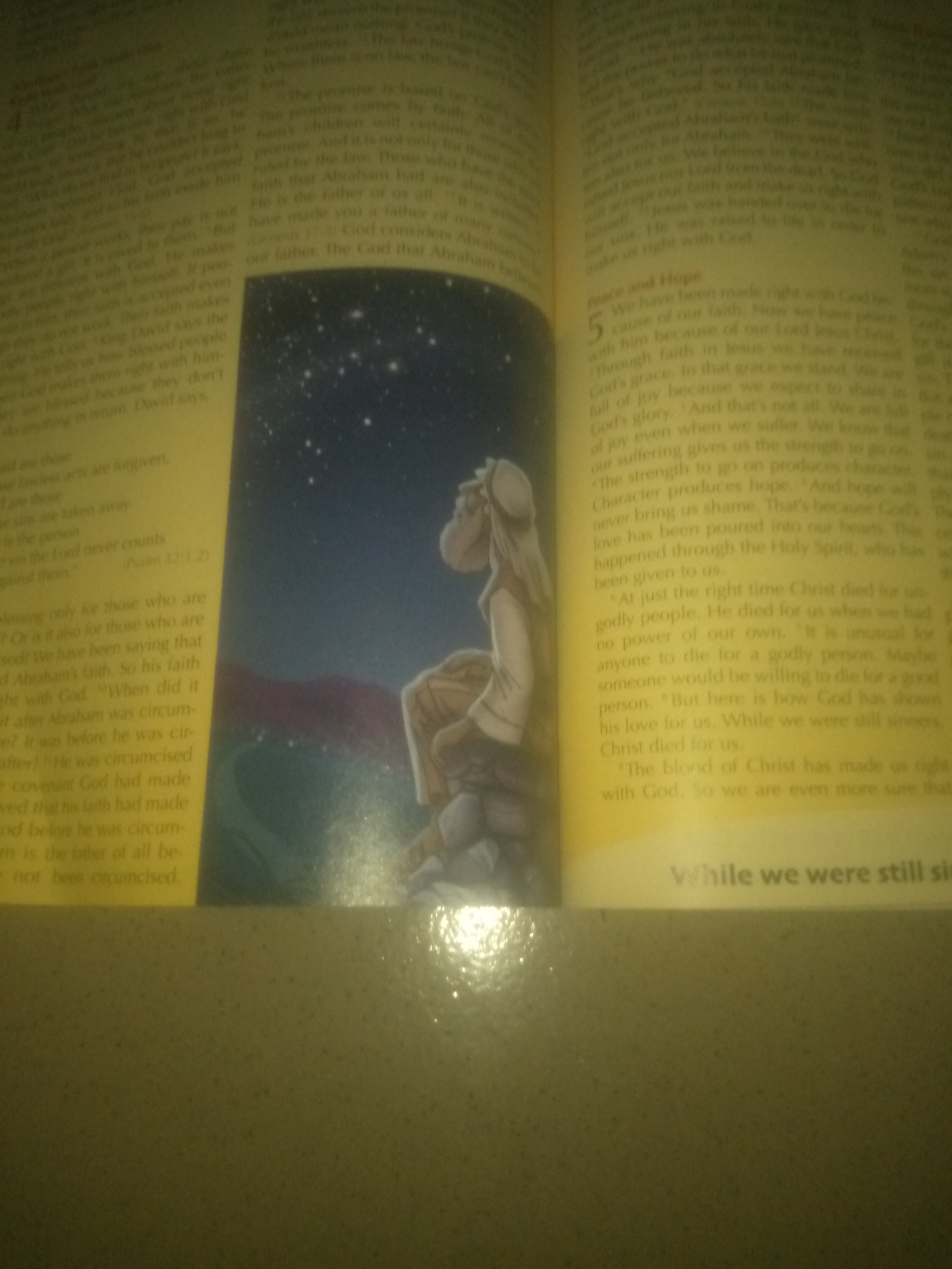 The image shows an open book with text and an illustration. The book is open to two pages, with the left page containing text and the right page containing text and an illustration. The illustration depicts a starry night sky with a figure sitting on rocks. The text on the left page is largely obscured by the illustration, but some phrases are visible, such as "the law never counts" and "against them." The right page contains text in paragraphs, with a numbered section labeled "Peace and Hope" and a subheading. Visible text includes "We have been made right with God," "through faith in Jesus we have received God's grace," "our suffering gives us the strength to go on," and "While we were still sin-ners, Christ died for us." The image is poorly lit, with a yellow tint and significant glare, making it difficult to read some of the text. There is no identifiable information within the image to suggest a location in Jauro Yinu, Nigeria.