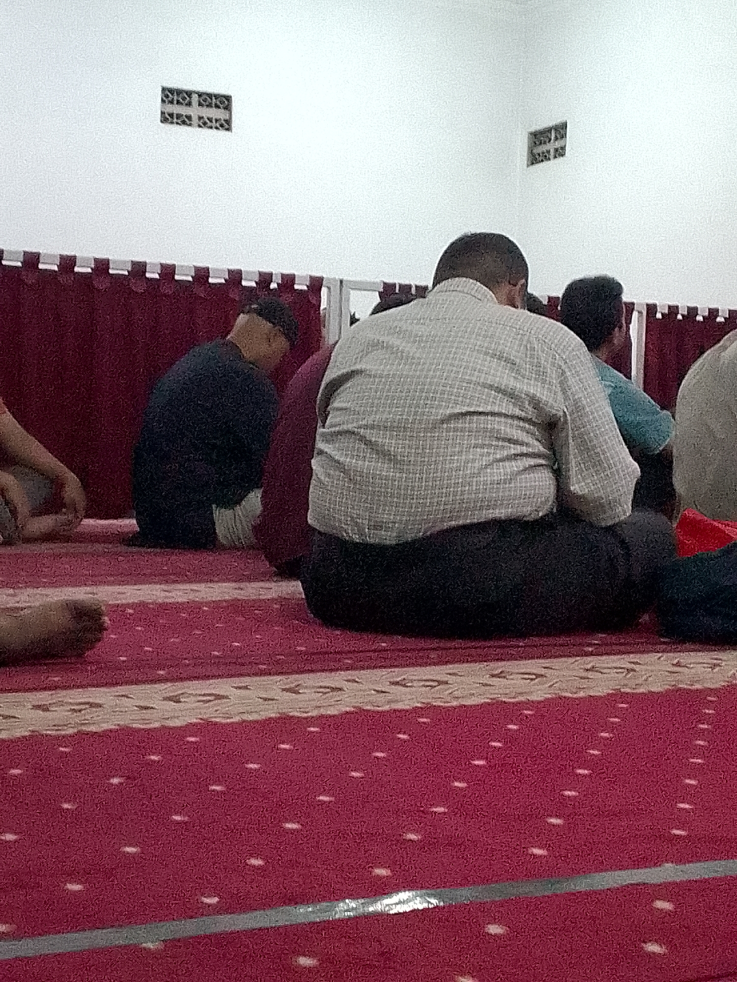 The image depicts a group of men sitting on a red carpet inside a building, likely a mosque, in Gunungmasigit, Indonesia. The men are seated in rows, facing away from the camera, with their backs to the viewer. They are dressed in traditional Islamic attire, including long robes and head coverings. Some men are wearing skullcaps, while others have their heads uncovered. The carpet has a patterned design with a central cream-colored stripe. The walls of the room are white and feature decorative elements. The overall atmosphere appears to be one of quiet contemplation or prayer.