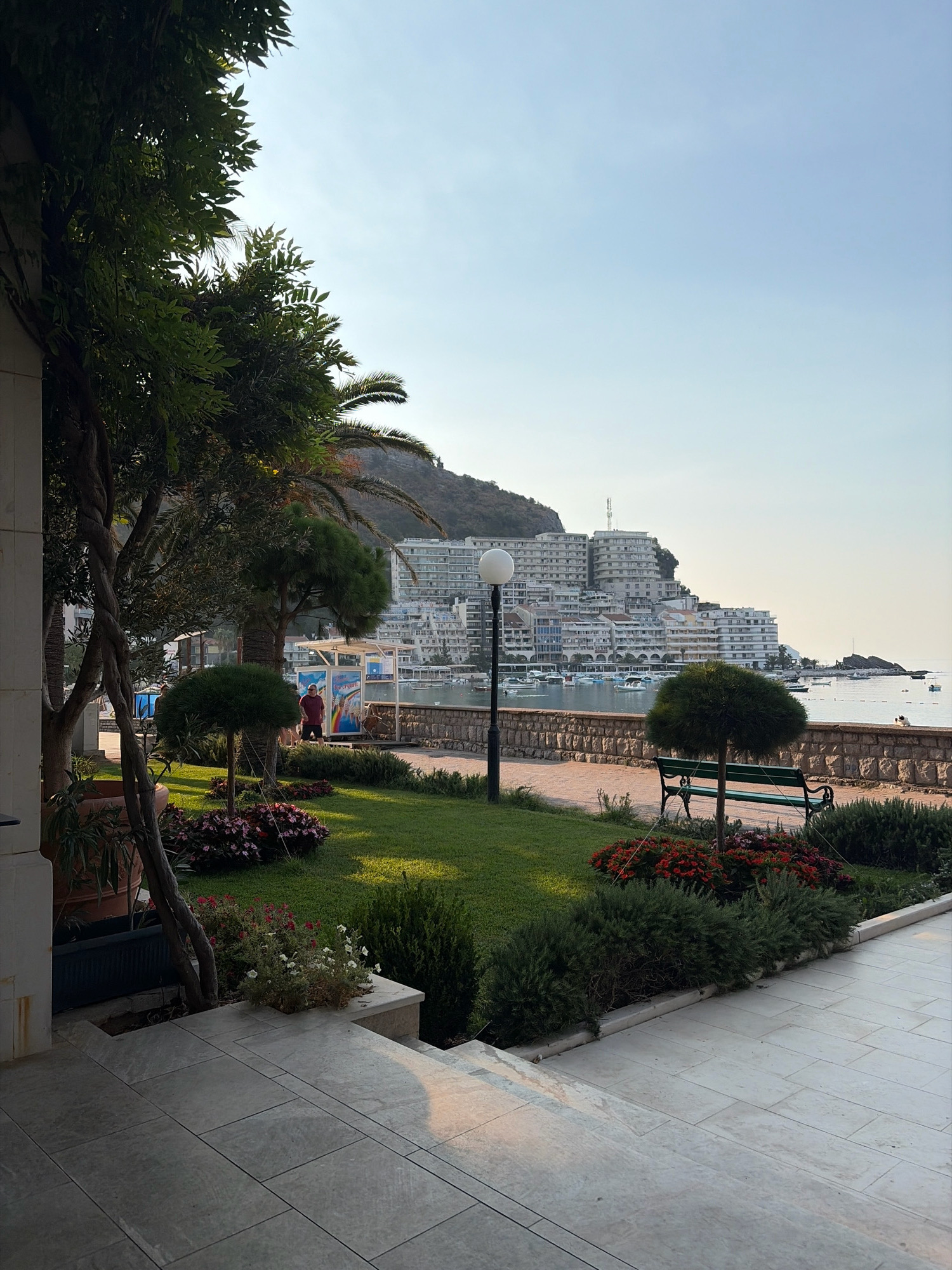 The image depicts a scenic view of the waterfront in Rafailovići, Montenegro. In the foreground, a paved walkway leads up a set of stairs, with lush green grass and well-maintained flowerbeds on either side. A bench is visible on the left side of the lawn, facing the water. In the middle ground, a stone wall separates the garden area from the harbor, where several boats are docked. A street lamp stands near the bench. In the background, a large complex of white buildings, characteristic of resort architecture, rises up the hillside. The sky is clear and blue, with the sun casting a warm light on the scene. A lone person in a red shirt is visible near a colorful structure on the waterfront. The overall impression is one of a tranquil and picturesque coastal town.