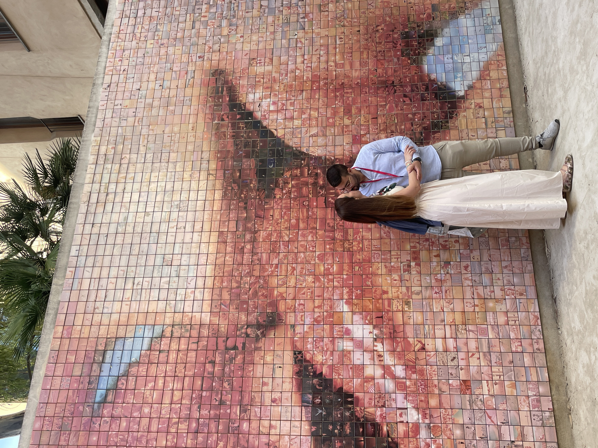 The image displays a couple intimately embracing in front of a large mosaic wall art located in Plaça d'Isidre Nonell, Barcelona, Spain. The mosaic, known as 'El Petó de la Llibertat' (The Kiss of Freedom), is composed of thousands of individual photographic tiles that collectively form a colossal image of two lips in a kiss. The artwork predominantly features warm tones of red, pink, and orange, with some tiles incorporating blue hues.
In the foreground, a man and a woman stand close, their heads tilted towards each other in an apparent kiss. The woman has long brown hair and wears a light-colored, floor-length maxi dress with patterned flat shoes. A dark blue jacket is draped over her left arm. The man, wearing glasses and a red lanyard, is dressed in a light blue button-up shirt, khaki trousers, and grey sneakers. His left arm is around the woman's lower back, and his right hand is clasped with her left hand.
The ground beneath the couple is a grey concrete pavement. Above the mosaic wall, vibrant green foliage, resembling palm fronds, and sections of a light-colored building are visible, framed against a bright sky, indicating an outdoor urban environment bathed in daylight.
