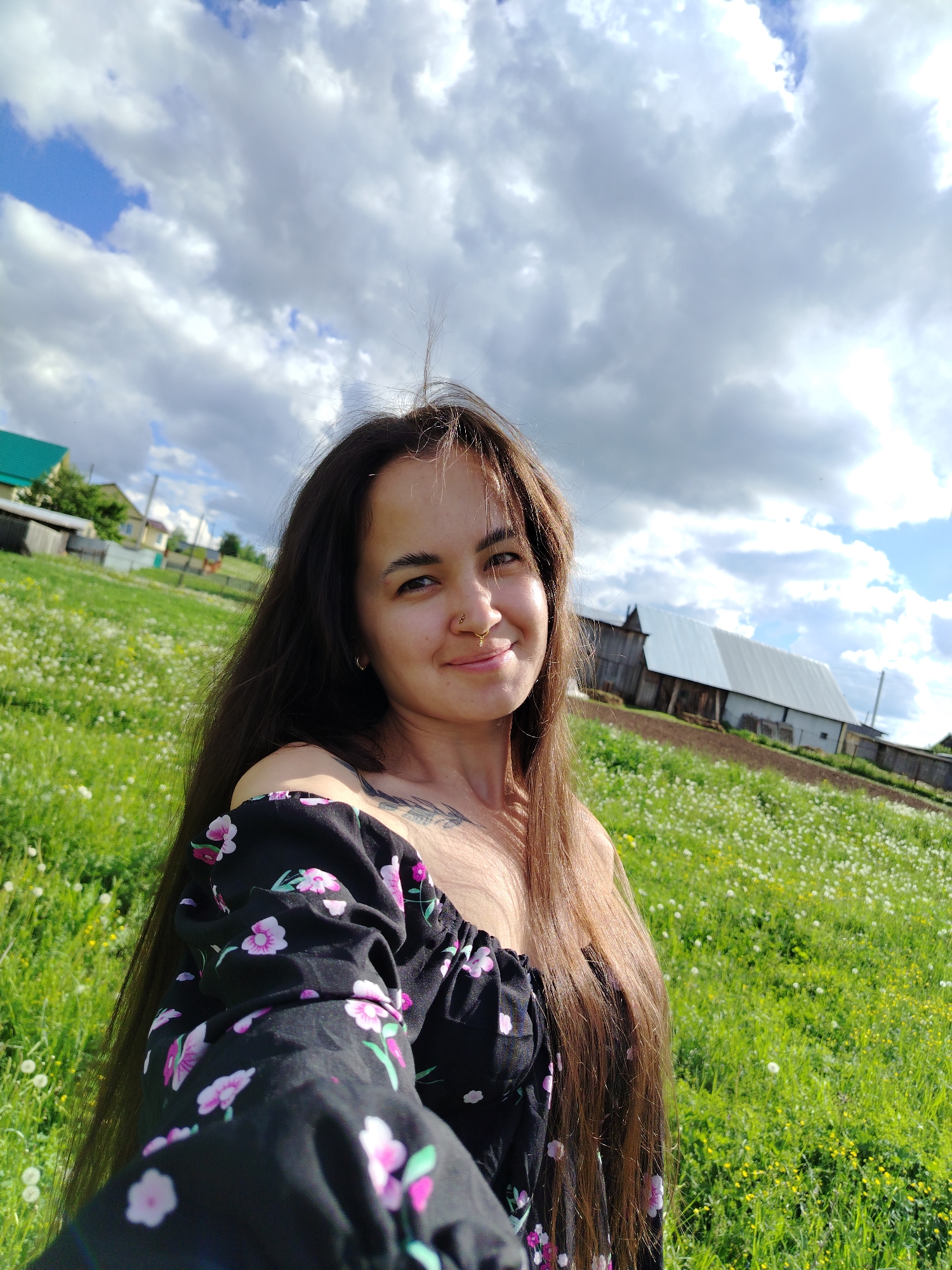 A young woman with long brown hair and a nose ring is taking a selfie. She is wearing a black off-the-shoulder dress with a floral pattern. There is a tattoo on her chest. She is standing in a grassy field with dandelions. In the background are buildings and a cloudy sky. The location is Гожан, Russia.