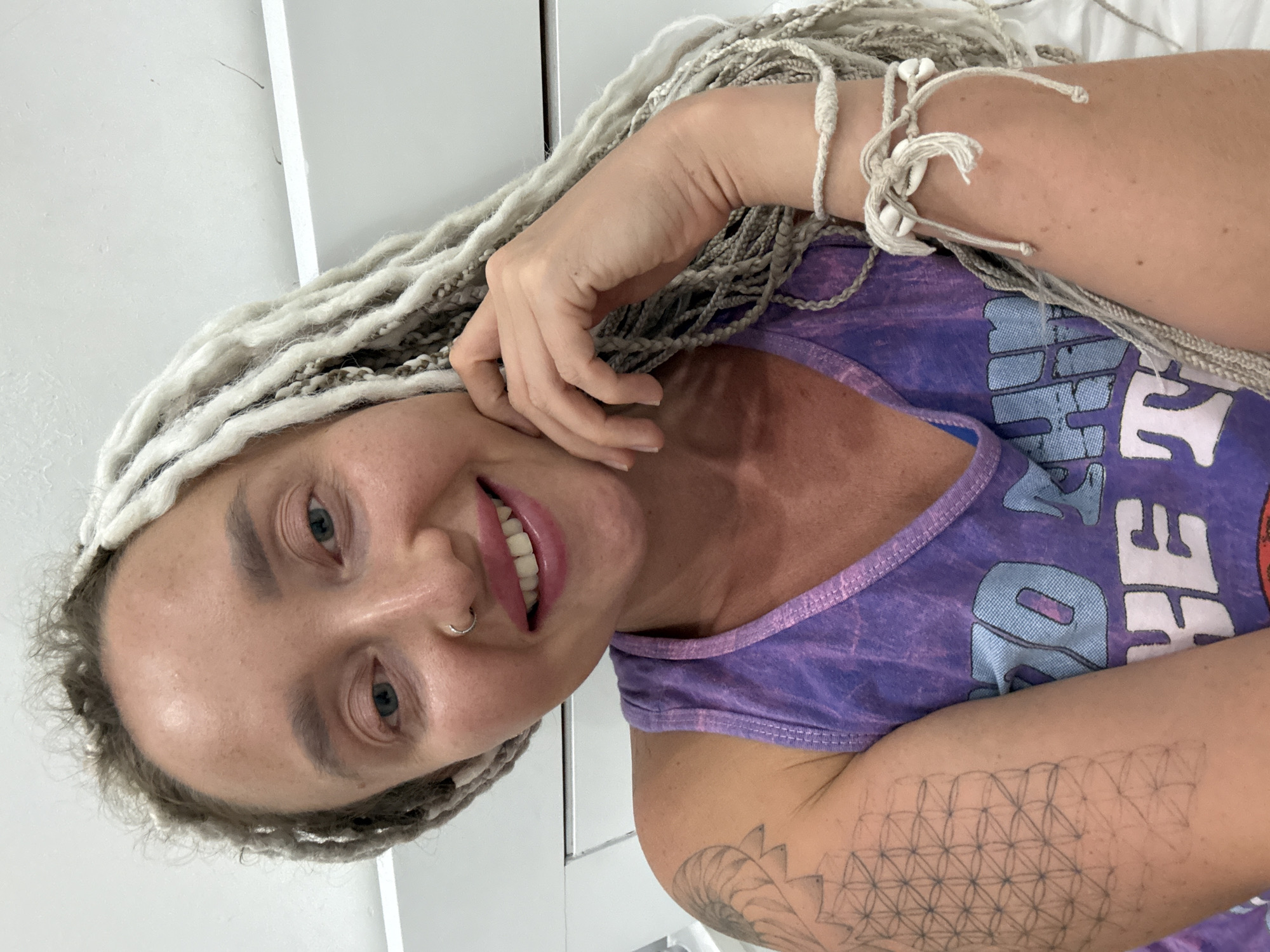 A close-up, waist-up shot shows a woman with light skin, a nose ring, and light-colored braided hair. She is wearing a purple tank top with blue and white lettering. Her right hand is resting on her chin, and her left arm is visible, showing a tattoo on her bicep and a bracelet on her wrist. The background consists of a white, paneled wall. The image does not contain any information about the city of Bac Nha Trang Ward, Vietnam, and therefore cannot be used to infer the location.