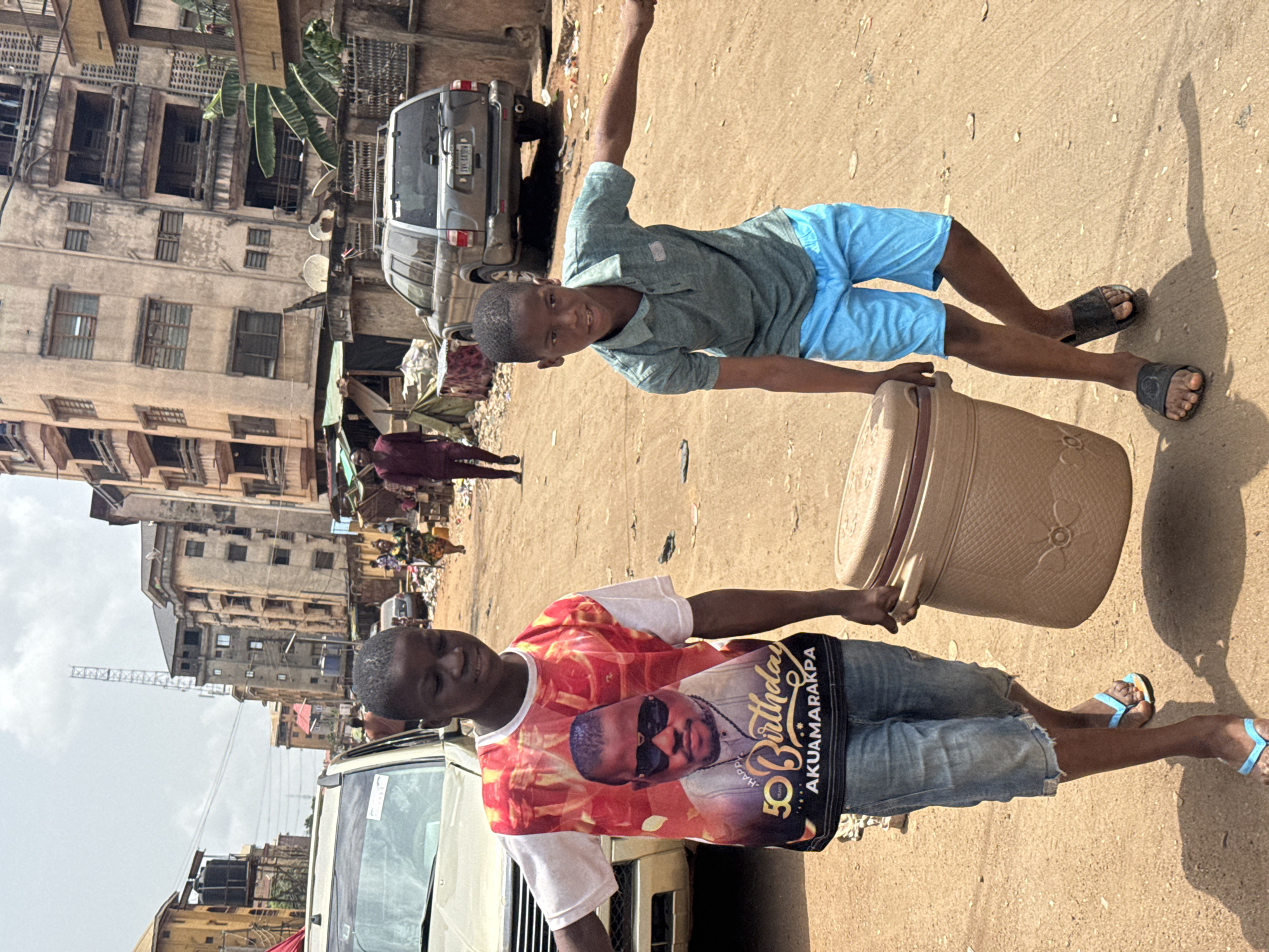 Two boys are outside in Onitsha, Nigeria. One is wearing a t-shirt with a photograph, blue shorts and sandals, carrying a plastic bucket. The second boy wears a gray shirt, blue shorts, and sandals, and is extending an arm. Two vehicles are parked nearby, along with a building. Other people are nearby.
