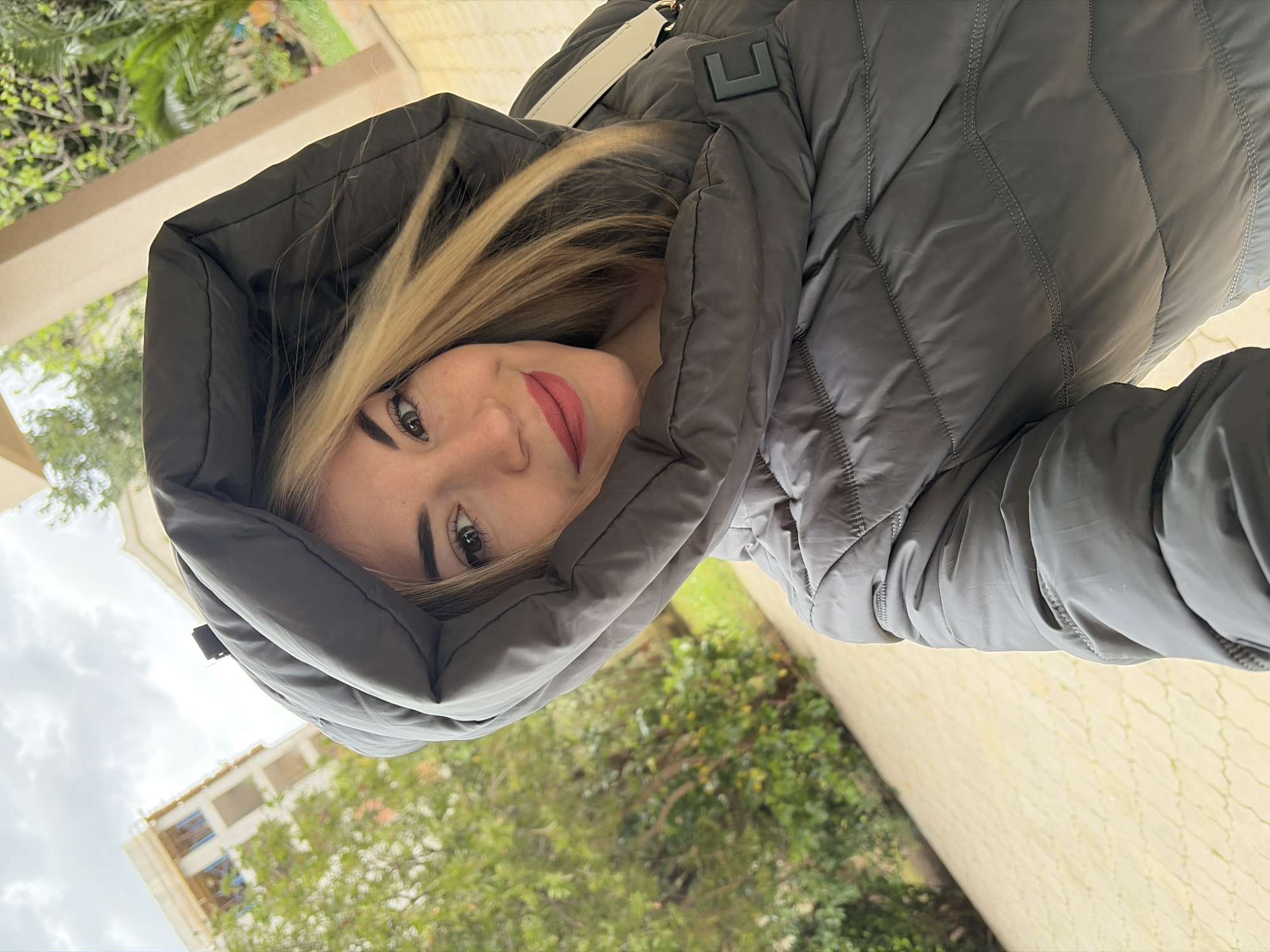 A woman with blonde hair and red lipstick is wearing a gray hooded puffer jacket. The hood is pulled up around her face. She is looking at the camera with a slight smile. The background shows green trees, building facades, and a cloudy sky. The location is Budva, Montenegro. The composition is taken from a slightly low angle, with the woman positioned off-center. The photo appears to have been taken outdoors during daylight hours.