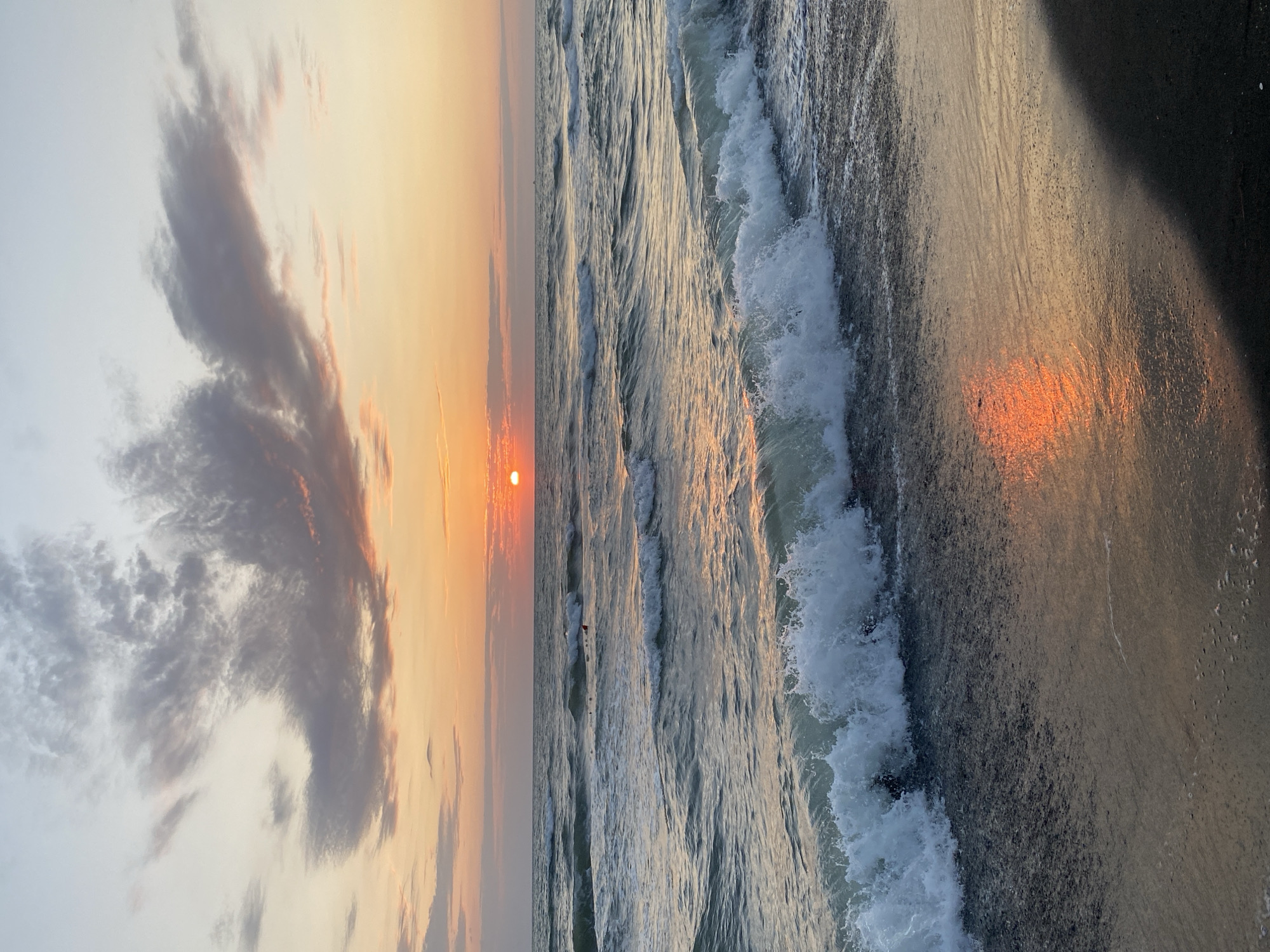 This is a photograph of a sunset over the Black Sea in Ureki, Georgia. The sun, a bright orange disk, is low on the horizon, casting a warm glow across the sky and water. The sky is filled with wispy clouds tinged with pink and orange. Waves are breaking on the shore, creating white foam. The beach in the foreground is dark sand. There are a few people in the water in the distance, appearing to be swimming or wading. The reflection of the sunset is visible on the wet sand.