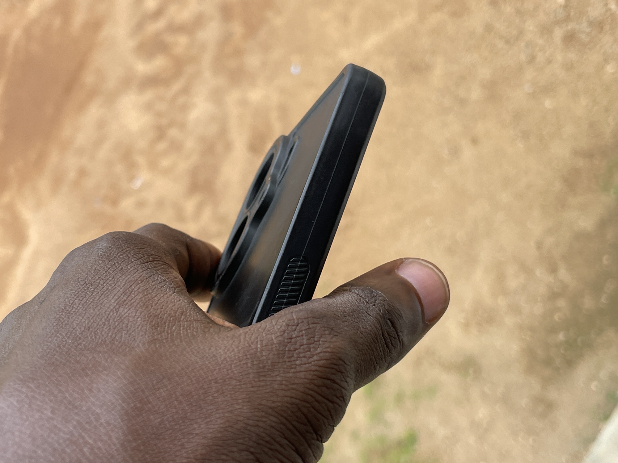 A close-up, oblique shot shows a dark-skinned hand holding a black phone case. The case is textured with a series of fine horizontal lines along its side, likely for grip. The background is a blurry expanse of light brown earth, suggestive of a dry, outdoor environment. The scene is devoid of any identifiable urban features, making it impossible to ascertain a specific location within Jos, Nigeria. There are no other individuals or significant objects present in the frame. The image focuses solely on the hand and the phone case.