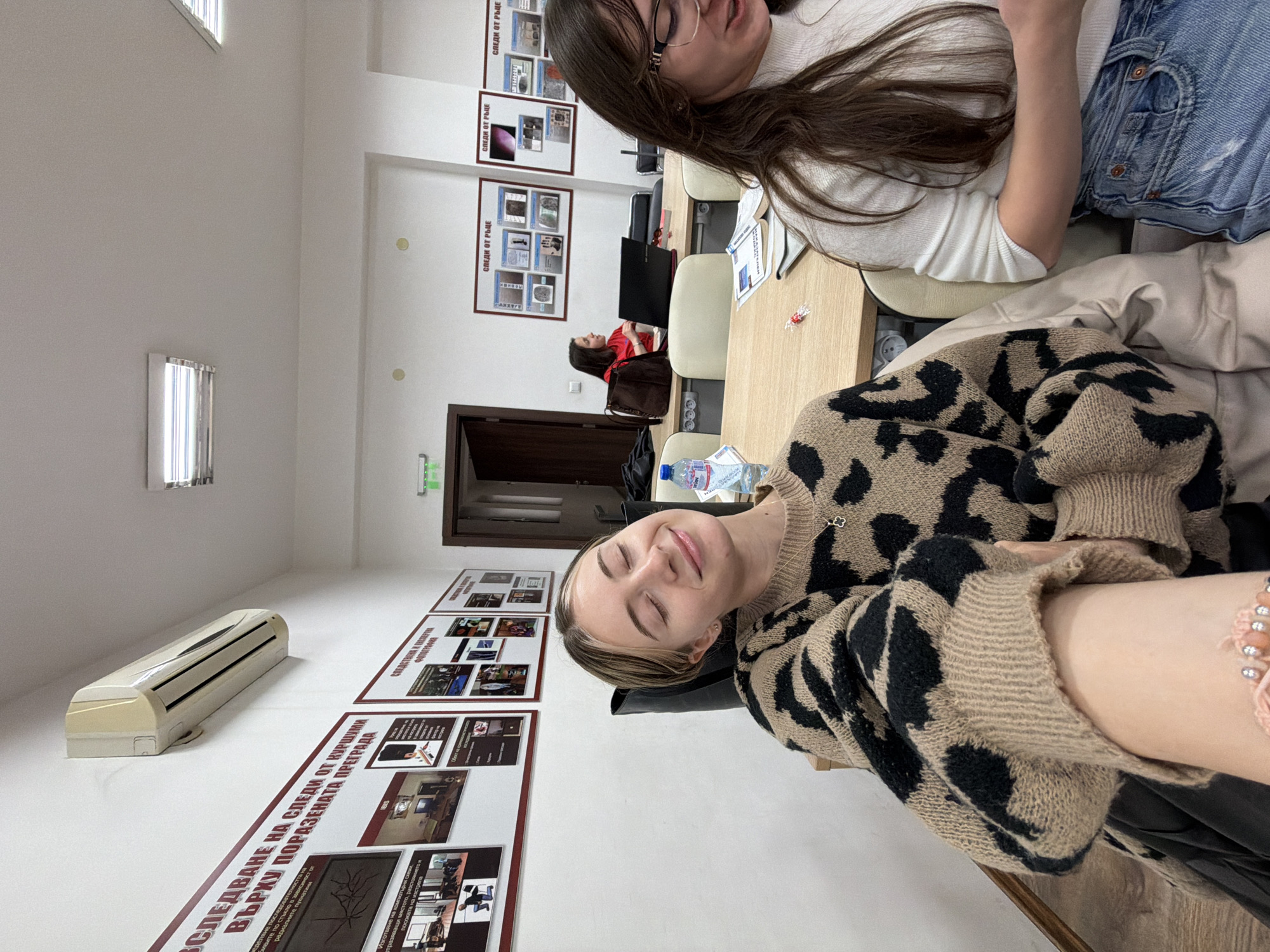 The image shows a room with posters on the wall, featuring the phrase "СЛЕДИ ОТ РЪЦЕ" and the text "СЛЕДВАНЕ НА СЛЕДИ ОТ КУРШУМИ ВЪРХУ ПОРАЗЕНАТА ПРЕГРАДА". There's an air conditioning unit installed on the wall. Two women are seated at a table, seemingly engaged in some activity, while another person is visible in the background. A bottle of water and other items are placed on the table. The photo appears to be taken in a classroom or office setting in Plovdiv, Bulgaria, based on the Cyrillic script in the posters.