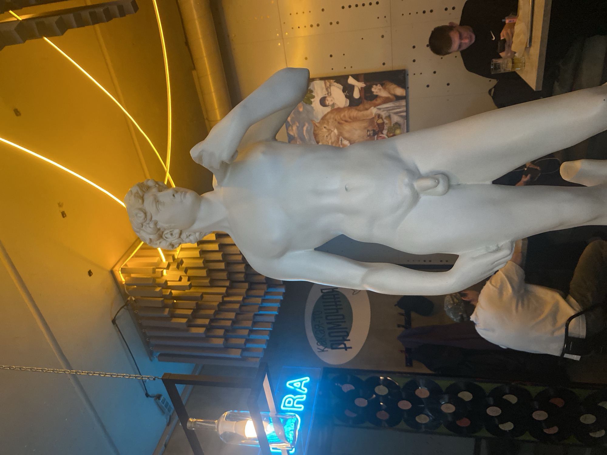 A statue of a nude male figure, reminiscent of Michelangelo's David, is centrally located in the frame. The statue is positioned in a space, potentially a cafe or bar in Krasnoyarsk, Russia, as evidenced by the Russian text "КАКАЯ-ТО РЮМОЧНАЯ" on a circular sign. Below the sign, there is a neon blue sign with the letter "RA" visible. A row of black vinyl records lines the bottom of the frame, partially obscured by furniture and people. In the background, a man in a black jacket is visible, seated at a table and using a mobile device. There is also another person in a white shirt. A framed picture of an oil painting hangs on the wall behind the statue. The environment is illuminated by warm yellow light, created by both exposed bulbs and neon lights.