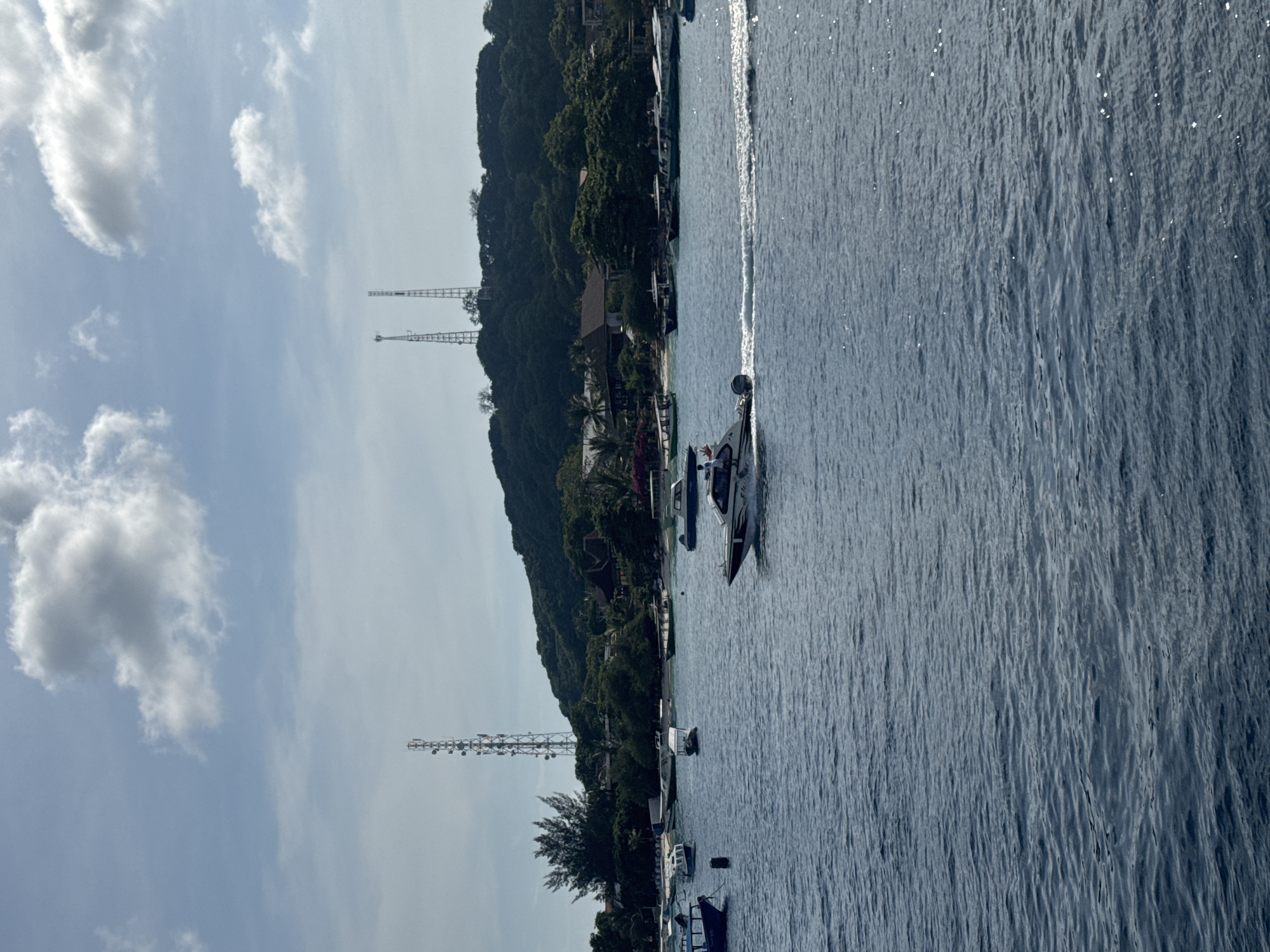 The image presents a scenic coastal view, captured in a vertical orientation. The left side of the frame is dominated by a partly cloudy sky, featuring several white and grey cumulus clouds against a light blue backdrop. Below the sky, stretching from the bottom to the top, is a heavily forested landmass, likely an island or a coastline, covered in dense green trees and tropical vegetation. Within this lush landscape, several structures are visible, including buildings with reddish-brown roofs nestled among the trees. Two prominent metal lattice communication towers stand tall on the hillside.
The right side of the image features a large body of water, likely the sea or ocean, with ripples reflecting bright sunlight across its surface. A white speedboat is actively cruising through the water, leaving a noticeable white wake in its path. A person aboard the speedboat can be seen raising an arm. Near the shoreline, which borders the forested landmass, several other smaller boats are either anchored or docked.
The scene takes place during the daytime, under sunny and partly cloudy weather conditions. The presence of tropical vegetation, boats, and the architecture suggests a coastal resort or populated island area, consistent with the location context of Indonesia. The activity depicted centers around maritime leisure and transport. No visible text is discernible in the image.