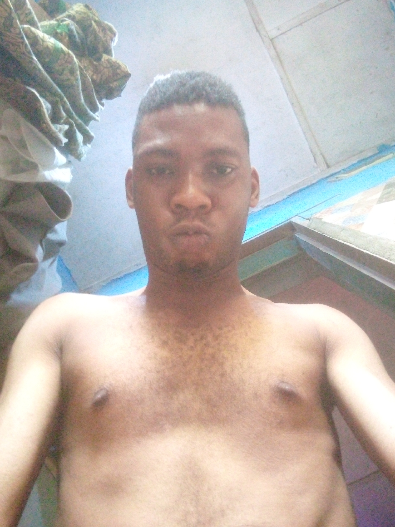 A selfie-style photograph features a shirtless dark-skinned man with short, dark hair. He is facing the camera with a slightly puckered expression. His upper body and arms are visible.
The background includes a pile of patterned fabric in the upper-left corner and a section of what appears to be a painted ceiling or wall, with blue and white colors.
The location is presumed to be Ikorodu, Nigeria, based on the prompt's instructions. No further details about the specific environment or location are visible.