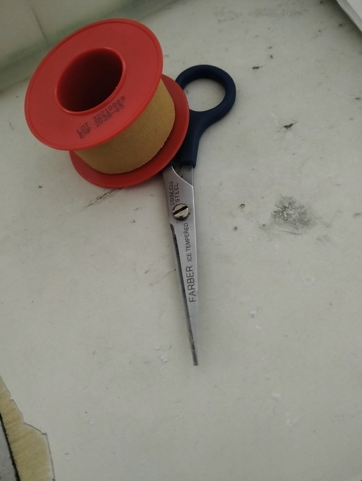 The image shows a roll of medical tape and a pair of scissors on a light-colored, textured surface. The medical tape has a red plastic dispenser and is partially unrolled, revealing a tan-colored adhesive bandage. Text printed on the tape dispenser reads "LOT 32210009 EXP 2028-05". The scissors have dark blue handles and a silver blade with "STAINLESS STEEL" and "FARBER ICE TEMPERED" printed on it. The surface appears to be a countertop or table, with some smudges and dark specks visible. The lighting suggests an indoor environment. There are no people or animals present in the image. The location is not identifiable as Malang, Indonesia based on the visual information provided.