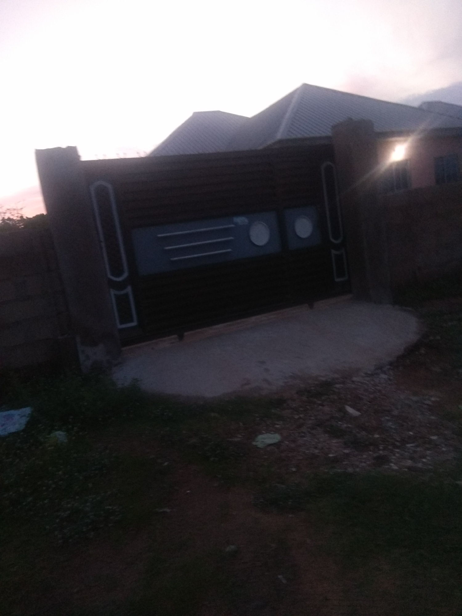 The image depicts a dark, horizontal gate in front of a building. The gate is made of dark horizontal slats with decorative white rectangular and circular elements. To the left of the gate is a short brick wall. To the right of the gate is a portion of the building, which has a dark roof and a window with light shining through it. In front of the gate is a patch of concrete. The ground in front of the gate and to the sides is covered with grass and dirt. The sky above is overcast and appears to be at dusk or dawn. The overall scene is dimly lit. This image appears to be a gate and building located in Jalingo, Nigeria.