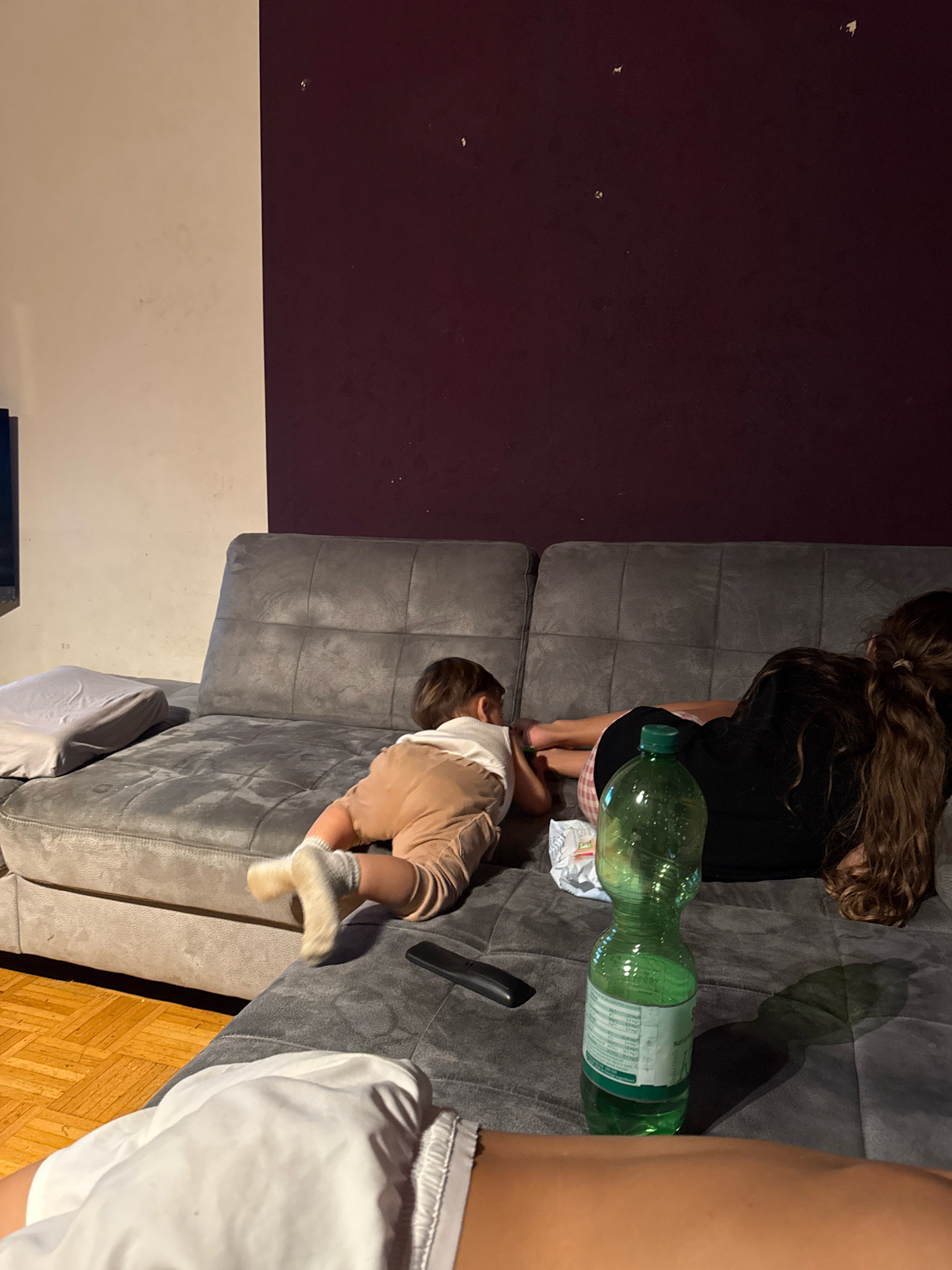 The image depicts a family relaxing on a sectional sofa in a dimly lit room. On the left side of the sofa, a young child is crawling on their hands and knees, wearing beige shorts and white socks. To the right of the child, an older individual, likely a sibling or parent, is lying down with their head resting on their arms. Their long, dark hair is visible, and they are wearing a black t-shirt and plaid shorts. In the foreground, a person's legs and torso are visible, wearing white shorts. A green plastic water bottle with a label is positioned on the sofa between the two individuals. The background consists of a two-toned wall, with the lower portion appearing to be cream-colored and the upper portion a deep reddish-purple. The floor is made of hardwood. There is no indication of the specific location within Überherrn, Germany.