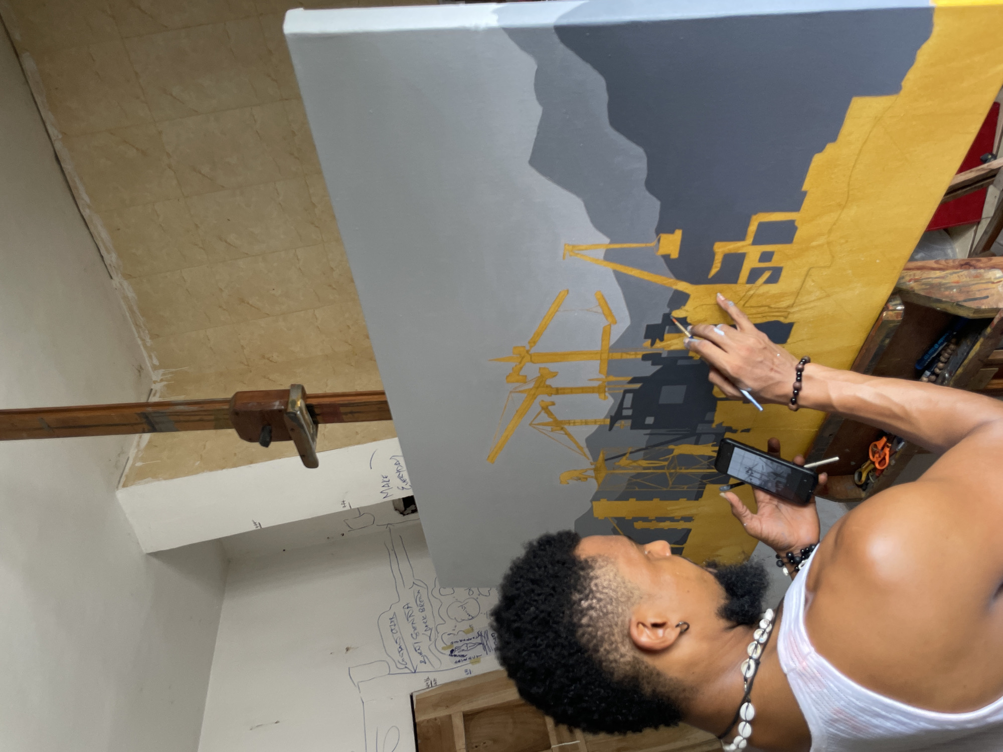 In an indoor setting in Ado, Nigeria, a man with dark skin, short curly black hair, a beard, and wearing a white tank top and shell necklace is painting on a large canvas. He holds a paintbrush in his right hand and a smartphone in his left. The canvas is predominantly gray and yellow, featuring silhouetted structures resembling industrial cranes and buildings against a mountain-like backdrop. The room has a tiled floor, white walls with sketches, and wooden supports for the canvas. The artwork is on an easel.