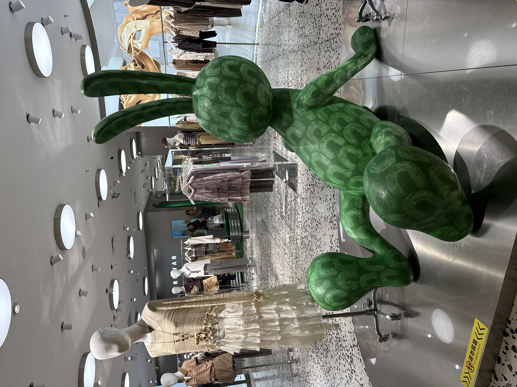 The image depicts the interior of a clothing store, likely a fashion boutique, in Huizhou, China. The store features a contemporary design with bright, even lighting from circular ceiling fixtures and spotlights. The floor is a speckled terrazzo material.
In the foreground, a large, bright green, stylized rabbit sculpture is prominently displayed. The sculpture is in a relaxed, seated position and has a textured surface with a pattern of green, curved lines. The rabbit is on a polished metal platform.
To the left of the sculpture, a mannequin is dressed in a bohemian-style outfit consisting of a crocheted top with fringe detailing and a long, tiered white lace skirt. Another mannequin, partially visible, is dressed in a brown and beige striped polo shirt and a skirt.
In the background, clothing racks filled with garments are visible, along with more mannequins displaying various outfits. The store's branding, "RIVAN," is visible on a sign above some of the clothing racks. Several individuals, appearing to be shoppers or staff, are present in the background, browsing or interacting within the store. A yellow sign with Chinese characters and English text, "NO CLAMPING ALLOWED," is visible near the bottom left corner of the image, on the metal platform.