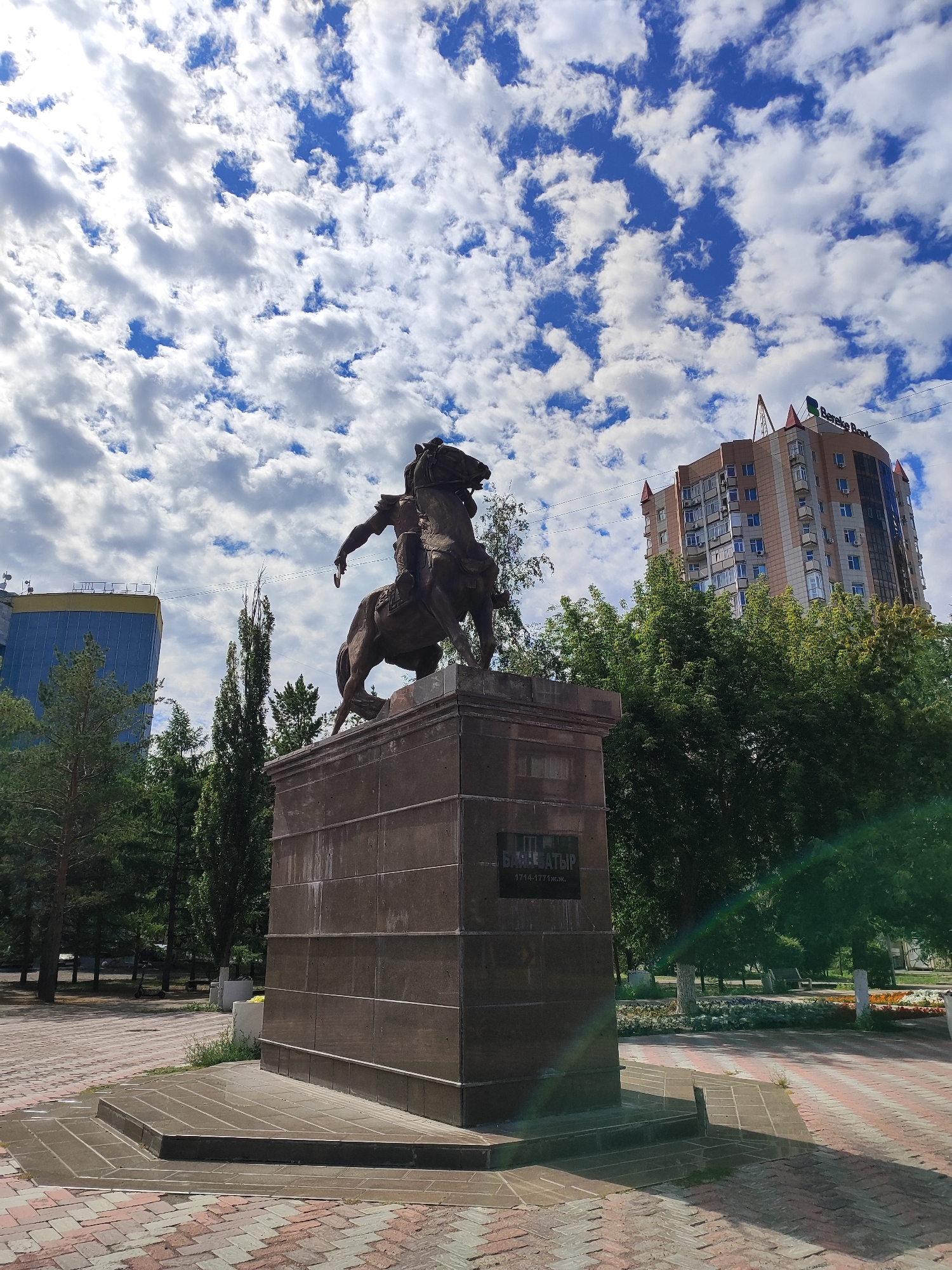 The image captures a monument of a man on horseback in a park-like setting in Pavlodar, Kazakhstan. The monument is positioned on a stone pedestal, and the man is depicted in a dynamic pose, suggesting action or command. The monument is inscribed with text that appears to be in Cyrillic script, with some of the letters and numbers, "1714-1771 ж.ж.", visible, indicating a historical figure or event.
In the background, modern city buildings are visible, including a curved, multi-story building with a "Bereke Bank" sign. Trees and greenery are abundant around the monument, indicating a well-maintained public space. The sky is filled with white cumulus clouds against a bright blue backdrop, suggesting a pleasant day. The ground is paved with interlocking bricks in a patterned design. The overall scene depicts a historical monument integrated into a contemporary urban environment, offering a contrast between the past and the present.