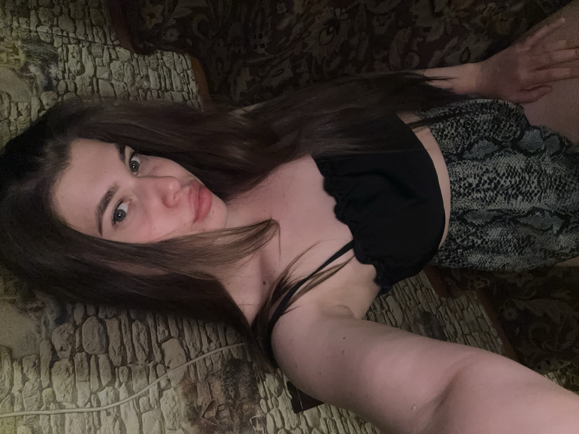 A young woman with long brown hair is lying on a couch or bed. She is wearing a black top and a patterned skirt or shorts. The background appears to be a wall or furniture with a textured surface, possibly resembling brick or stone. The image is a close-up selfie-style photograph. The location in the city of Мещёры, Russia is not identifiable based on the visual information provided in the image.