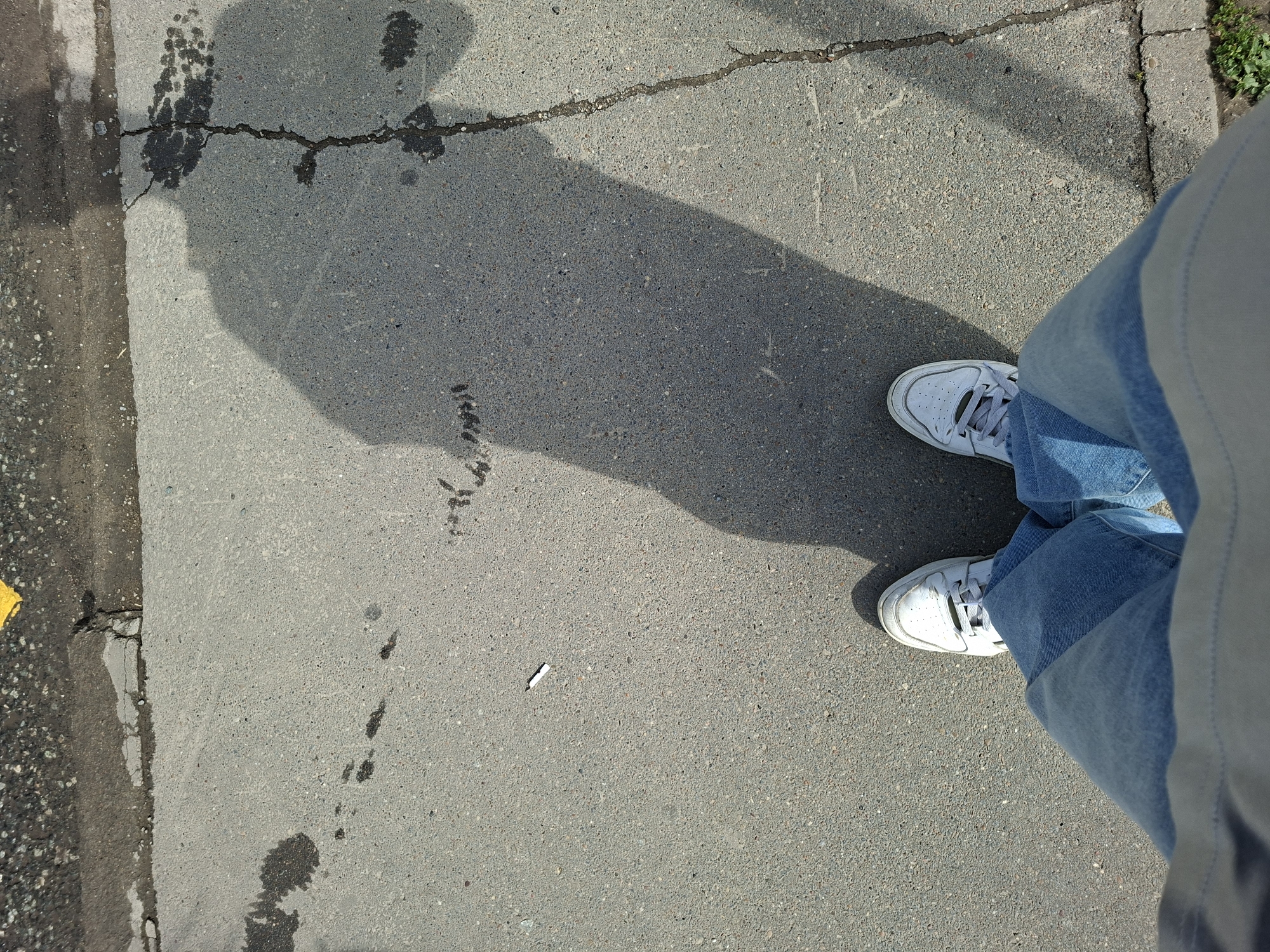 The media file shows a first-person perspective of someone standing on a cracked asphalt surface, likely a sidewalk or pathway. The subject is wearing white sneakers and light-wash denim jeans. A shadow from an unseen object or person is cast across the asphalt, suggesting it's daytime. There are several dark splatters and some footprints on the ground, indicating recent moisture or spills. A discarded cigarette butt lies on the ground near the center of the frame. The lighting suggests it is a sunny day. The background hints at an urban or suburban environment with the edge of a road and some greenery visible on the right side. The overall scene conveys a casual, everyday moment.