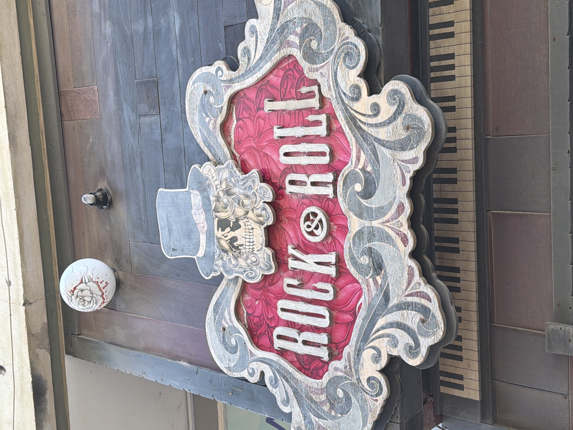 The image shows a close-up of a decorative sign that reads "ROCK & ROLL" in stylized, vertically stacked letters. The sign is set within an ornate frame, featuring scrollwork and a skull wearing a top hat. The background of the sign is a red floral pattern. Adjacent to the sign is a painted keyboard graphic and a wooden paneled wall with a white doorknob featuring a flower pattern. The location is Alexandroupolis, Greece.