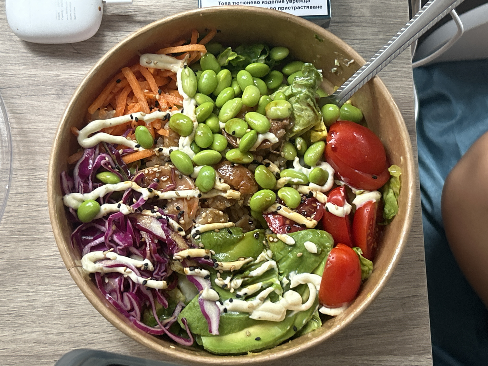 A close-up, overhead shot shows a poke bowl served in a disposable cardboard container placed on a wooden table. The bowl is generously filled with a variety of ingredients. At the top, shredded carrots are visible, interspersed with edamame beans. A creamy white sauce is drizzled over the contents. In the center, there are pieces of what appears to be cooked meat or fish, along with more edamame and lettuce. The left side of the bowl features shredded purple cabbage and sliced avocado, also adorned with the white sauce and sprinkled with sesame seeds. The right side of the bowl contains halved cherry tomatoes and more lettuce. A fork with a textured handle is partially submerged in the bowl, suggesting the food is being eaten. In the background, a white electronic device and a corner of a blue cloth are visible. The scene appears to be indoors, possibly in a cafe or at home, during daylight hours. Text is partially visible on a white object in the upper background, and some of the text is in Cyrillic. The overall impression is that of a healthy and appealing meal being enjoyed.