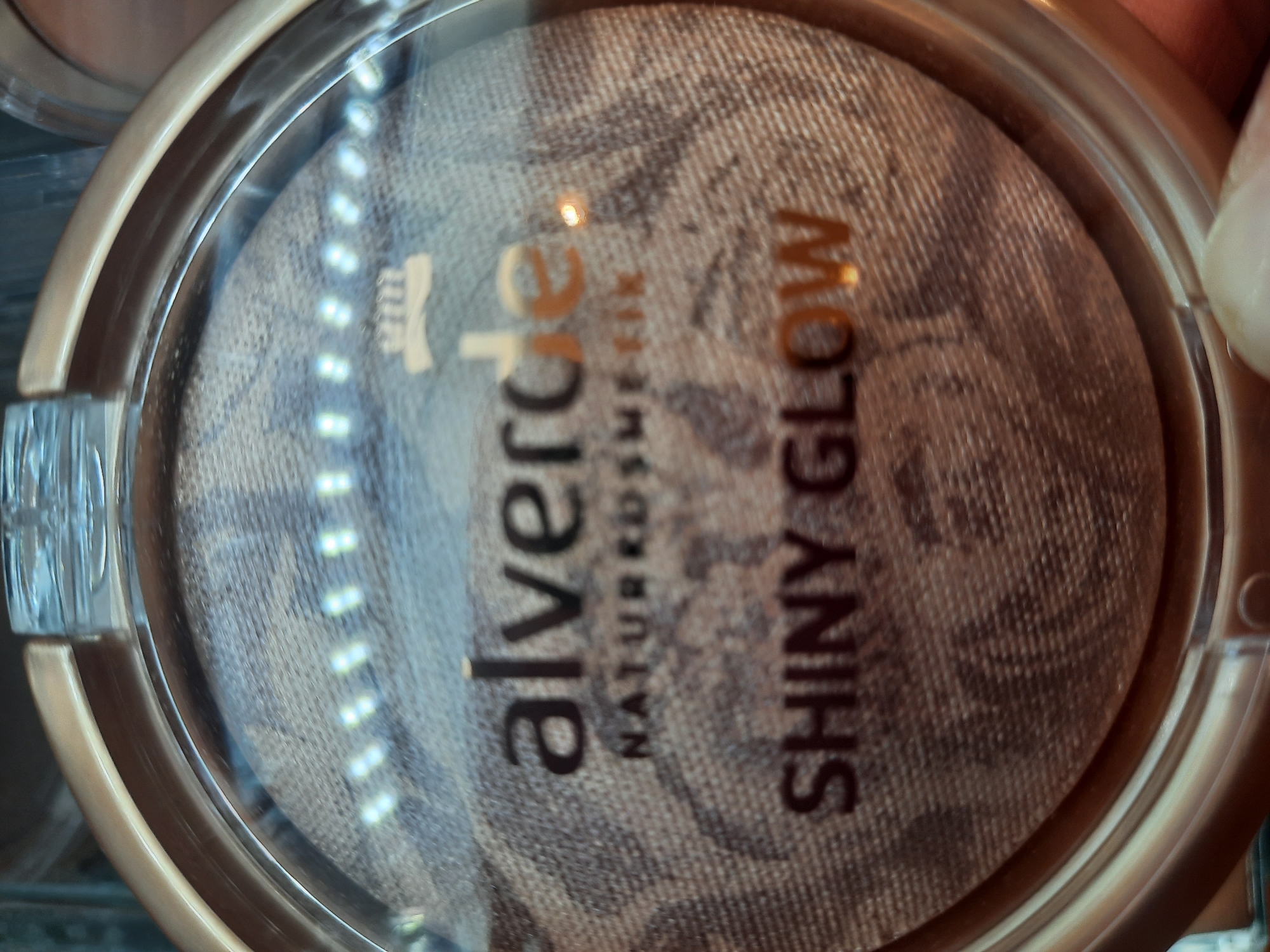 The image shows a close-up, slightly blurry view of a makeup compact. The compact is round with a gold-toned outer casing and a transparent lid. Inside the compact, there is a pressed powder product with a marbled pattern. Text is visible on the product and the packaging. The top line of text reads "alverde", with "NATURKOSMETIK" below it. Below that, in larger letters, is "SHINY GLOW". A small, stylized graphic of what appears to be a laurel wreath or crown is visible above the "alverde" text. The background is out of focus but suggests an indoor environment with some lighting elements visible as bright white dots. The location of this image in Rosenheim, Germany is not ascertainable from the visual information provided. There are no people or actions depicted in the image.