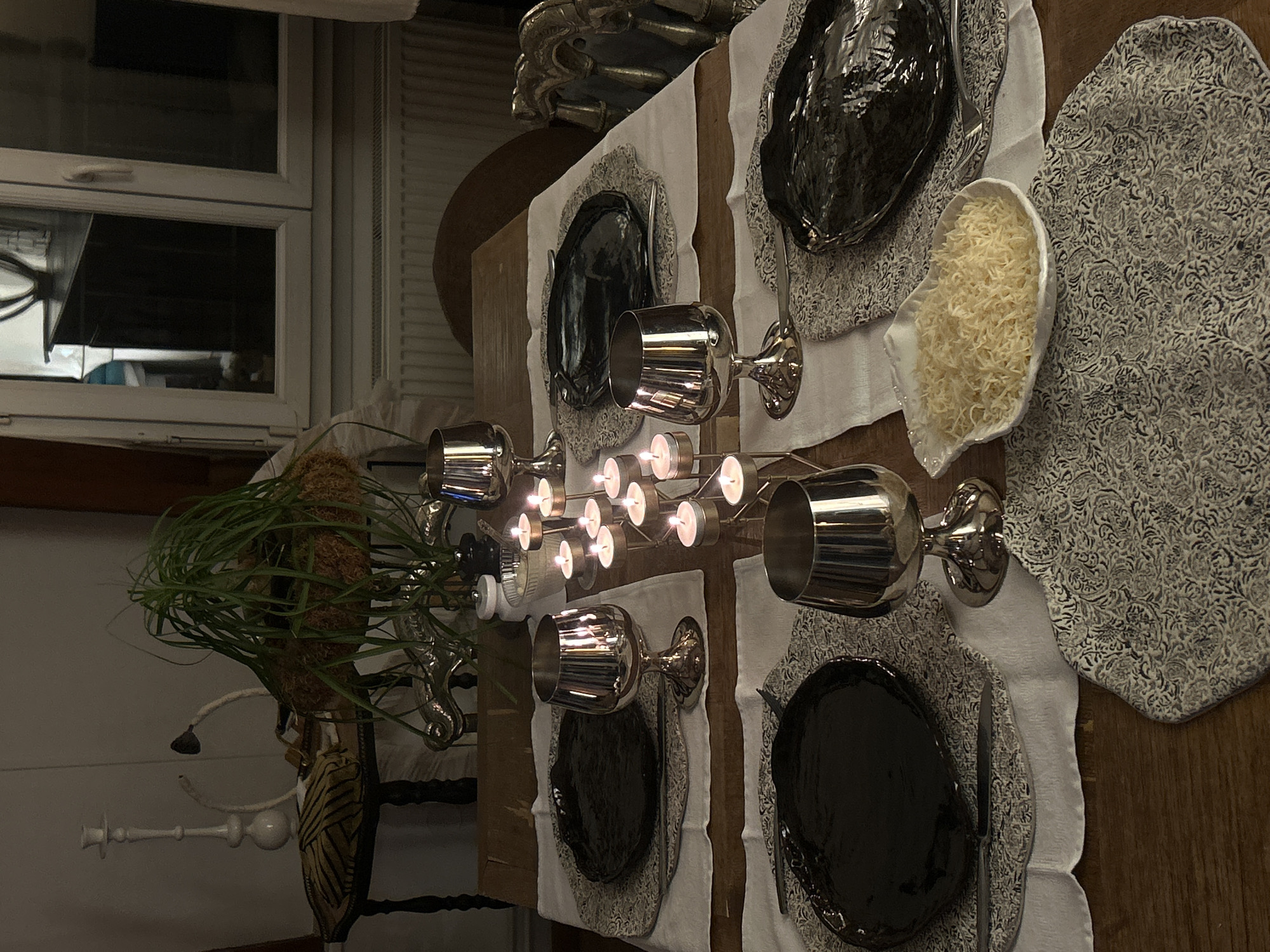 The image depicts a formally set dining table in an indoor environment. The table is wooden, covered with white runners and patterned placemats in shades of gray. Each place setting includes a dark, possibly black, decorative plate and silverware. There are also silver goblets, and a white bowl filled with a light-colored food item, possibly pasta or rice. A candelabra with lit candles is centrally placed on the table. In the background, there is a window and what appears to be decorative furniture, including a plant.