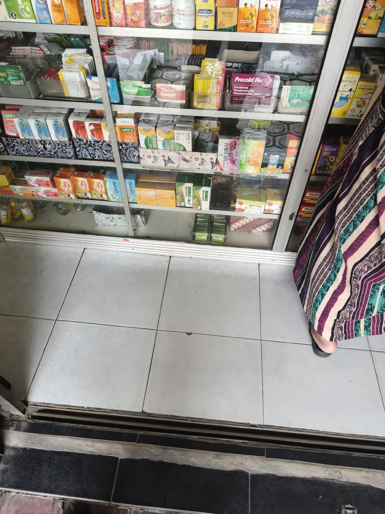 This is a medium shot of the interior of a pharmacy shelf in Sidorejo, Indonesia. The shelves are stocked with various health and personal care products, including boxes of medication, vitamins, and toiletries. The products are neatly arranged, with some items in their original packaging and others displayed individually. The lighting is bright and even, suggesting an indoor environment. A person's feet and the lower part of their colorful striped skirt are visible on the right side of the image, indicating they are standing in front of the shelf. The floor is tiled with white squares.