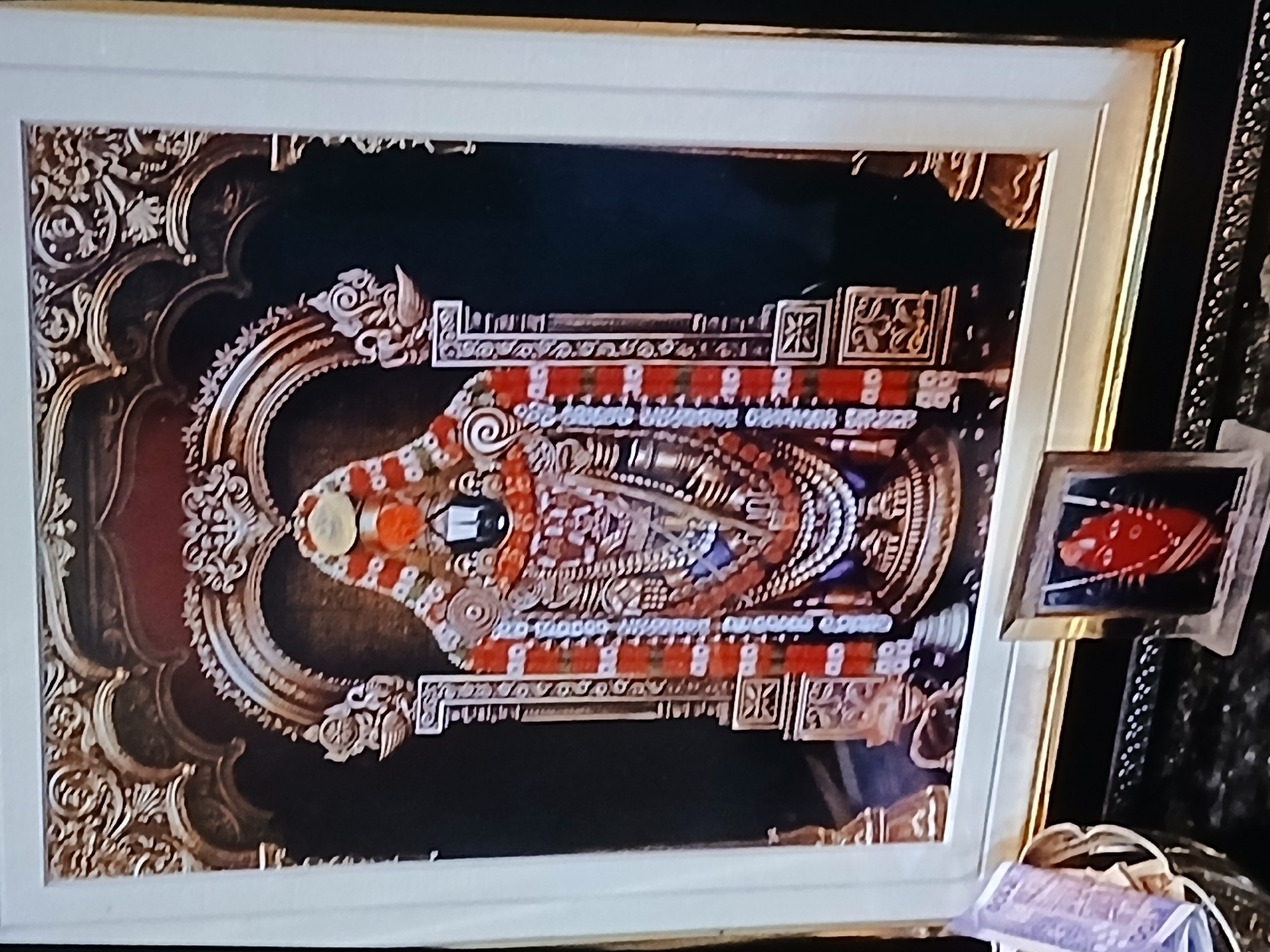 The image shows a framed artwork depicting a deity, likely Vishnu or a related avatar, adorned with elaborate jewelry and floral garlands. The artwork is framed with an ornate, gold-colored pattern on a dark background. The overall style appears traditional South Indian. Adjacent to the framed artwork is a smaller, framed image featuring a red object with white stripes, possibly a fish or a similar motif. The environment suggests a Hindu home or temple interior. Considering the artistic style and the deity depicted, Bengaluru, India, is a plausible location due to its large Hindu population and numerous temples.