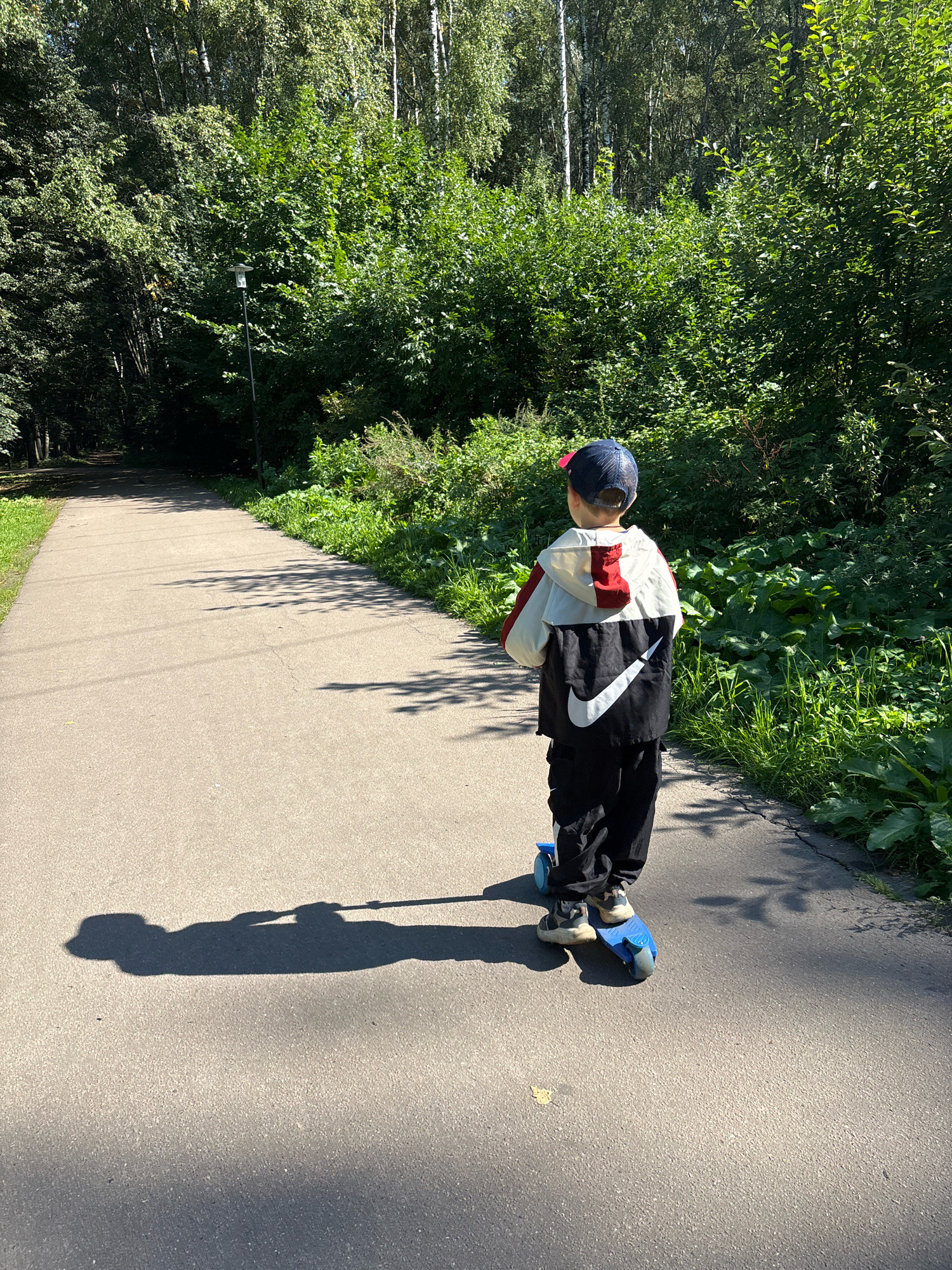 In Rusanovo, Russia, a young boy, seen from the rear, rides a blue scooter along a paved park path. He wears a blue baseball cap with a red brim, a white and red jacket with a large white Nike swoosh logo on the back, and black pants. The path is bordered by lush green trees and foliage on both sides, with sunlight casting dappled shadows. The scene appears to be a sunny day in a park.