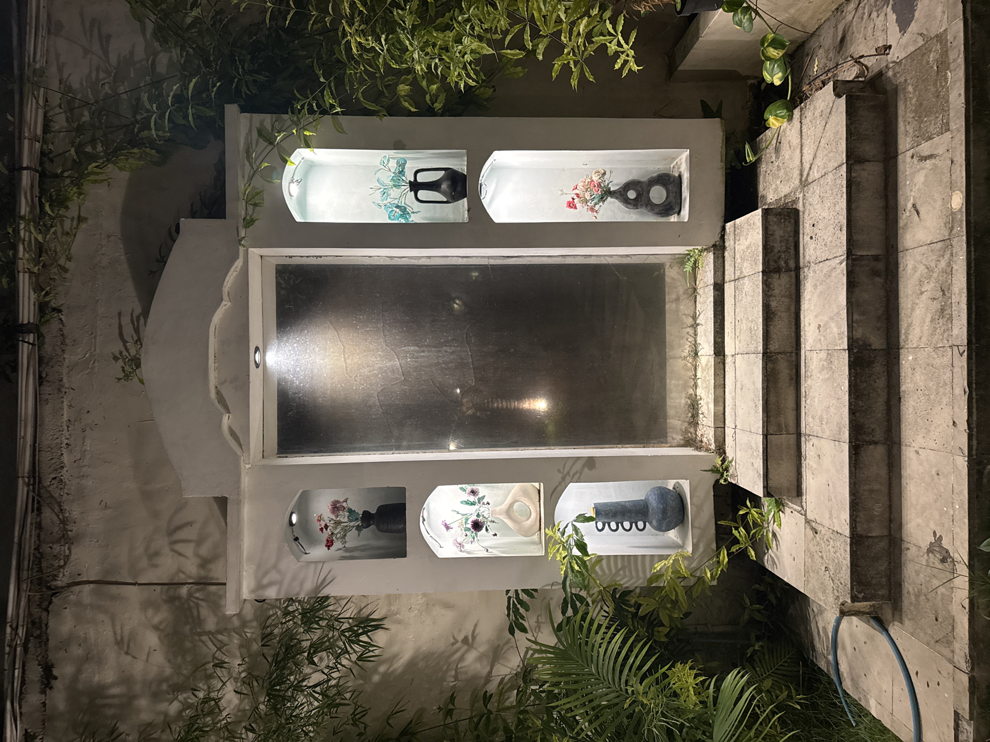This image depicts an outdoor entryway at night in Kuta Selatan, Indonesia. The entryway features a central glass door flanked by a series of recessed niches on either side. Each niche contains a decorative vase, some with artificial flowers. The niches are illuminated from above. In front of the entryway, a set of concrete steps leads up from a tiled ground level. Greenery, including palm fronds and vines, surrounds the structure. The overall impression is of an illuminated architectural feature set against a dark, natural backdrop.