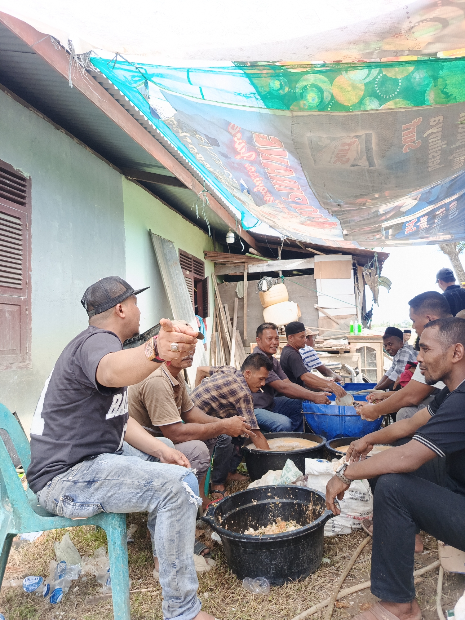 In Sangso, Indonesia, a group of men are gathered outdoors, seated on the ground and chairs under a makeshift tarp canopy. The canopy, made of assorted fabrics and banners, provides shade from the sun. Several men are actively engaged in food preparation, with some reaching into large plastic basins filled with what appears to be chopped food ingredients. One man in a black t-shirt and cap is gesturing towards the group, while others are focused on their tasks. There are several large black and blue basins, along with bags of what might be rice or flour. The environment is rustic, with a light blue-colored building in the background and scattered debris on the ground, suggesting a casual outdoor gathering or event.