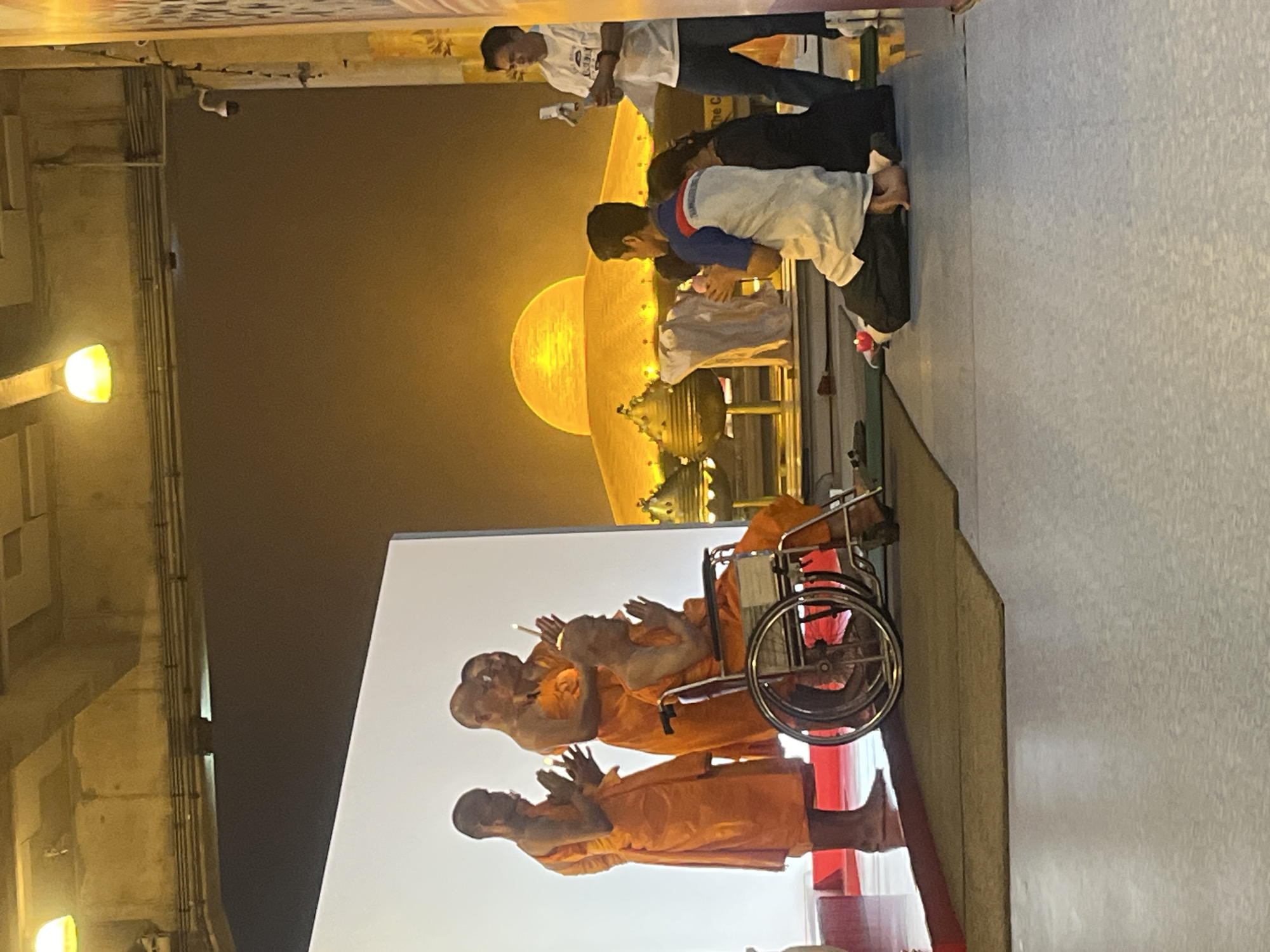 The image depicts an outdoor event at the Phra Pathom Chedi in Tha Khlong Town Municipality, Thailand. Several Buddhist monks, dressed in orange robes, are present. One monk is seated in a wheelchair and appears to be receiving a blessing or participating in a ceremony. Other monks are standing, holding lit candles and with their hands in prayer positions. In the background, the iconic Phra Pathom Chedi is visible, illuminated by a golden light, suggesting it is either dawn or dusk, or the structure is artificially lit. Laypeople are also present, with some kneeling and others standing. The environment appears to be a temple or religious complex, with paved ground and an overhead structure suggesting a covered area. The overall scene conveys a sense of solemnity and religious observance.