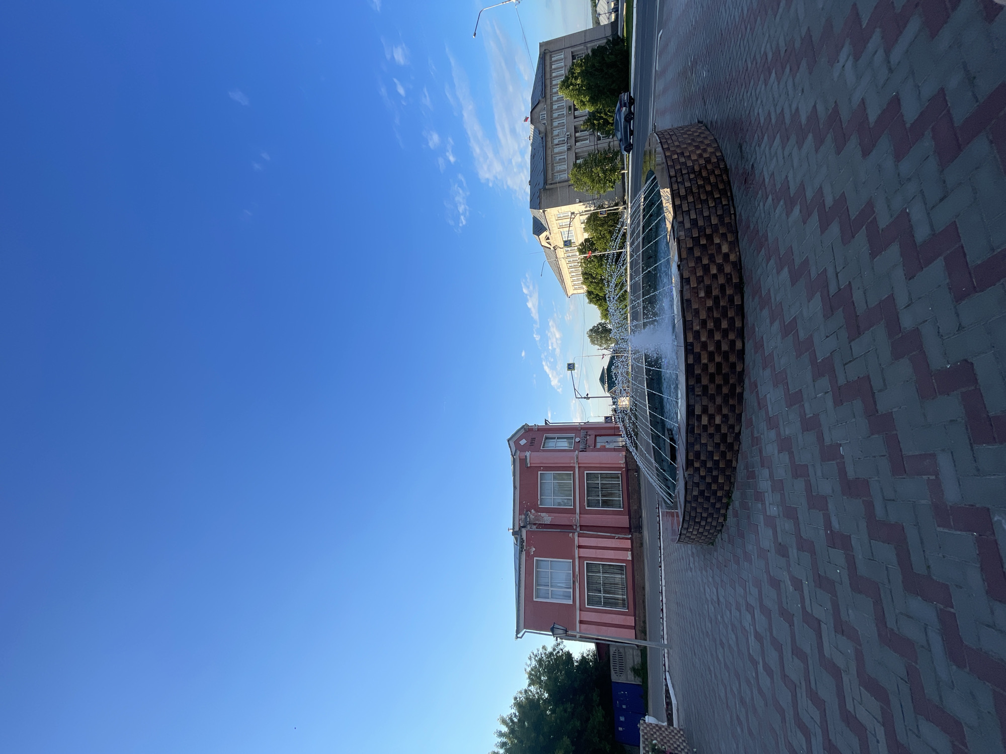 This is a photograph of a street scene in Krasnye Baki, Russia, featuring a circular fountain in the foreground. The fountain has a brick-like base and is actively spraying water upwards, creating a mist. The ground surrounding the fountain is paved with interlocking bricks laid in a herringbone pattern.
In the background, on the left, is a two-story building painted pink with white trim. It has multiple windows with curtains. To the right of the pink building, across a road, is a larger, lighter-colored building with a gray roof, multiple windows, and a flag flying from the roof. Trees are visible between the buildings and along the sidewalk. A dark-colored car is driving on the road. The sky is a clear blue with a few wispy clouds. A street light is visible to the left of the pink building.