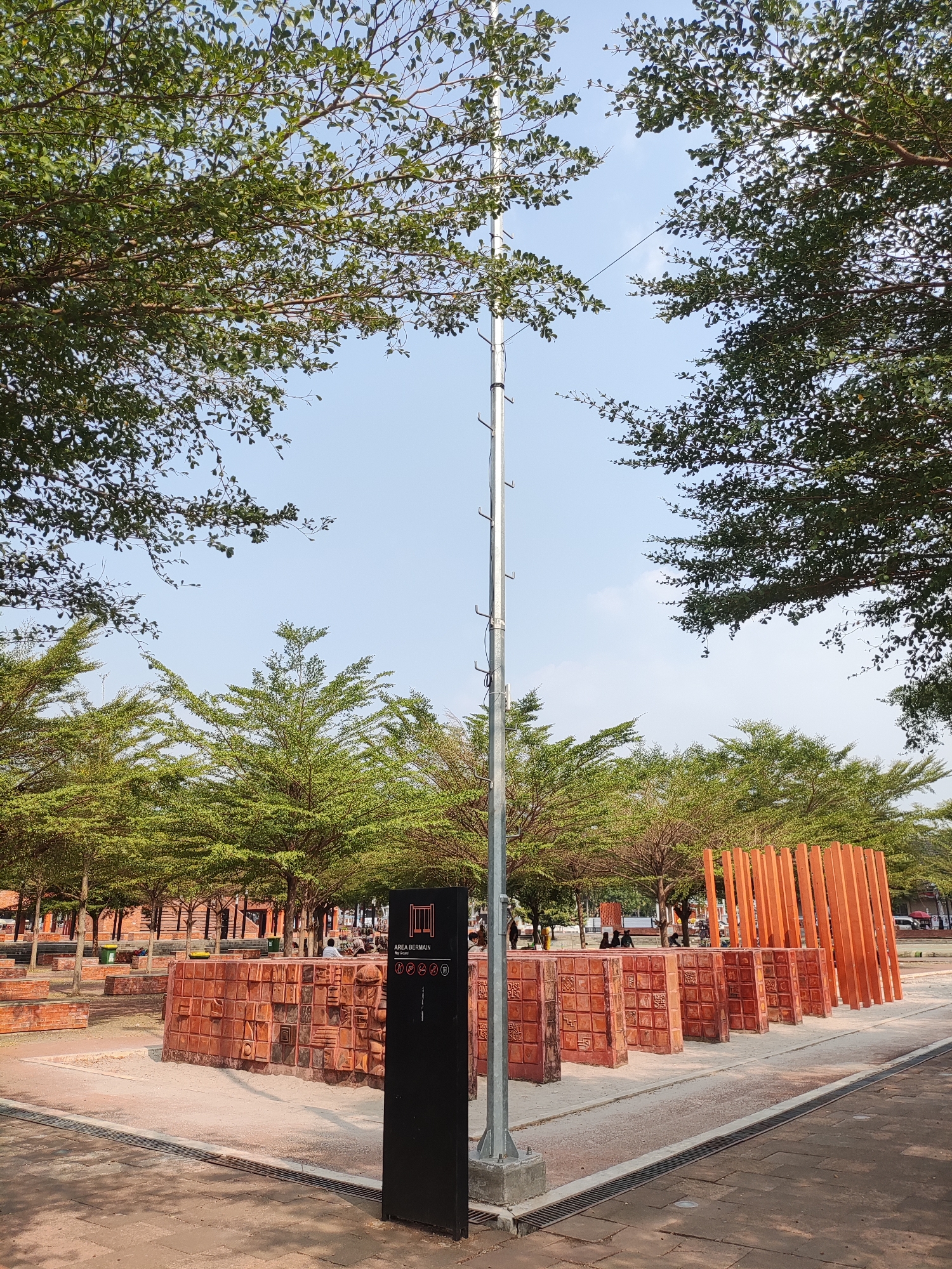 This is a full-shot, eye-level, vertical photograph of a public outdoor area. The foreground shows a paved walkway with drainage grates. A tall, silver, pole-like structure with multiple hooks is positioned on the left side of the frame, topped by thin wires extending to the right. To the right of this pole is a tall, black sign with white text and icons, including the words "AREA BERMAIN". Behind the sign is a circular, low-walled structure constructed from red, patterned bricks, forming a seating or display area. Several identical, orange, vertical wooden planks stand behind the brick structure, angled slightly.
In the middle ground, a park-like setting is visible with numerous trees with sparse, green foliage, casting shadows on the ground. The trees are arranged in rows, creating a canopy effect. Several people are seated in the background, some near the brick structure and others further back. Brick buildings are visible behind the trees on the left.
The background features a clear, pale blue sky with a few wispy white clouds. The overall environment suggests a well-maintained public space, possibly a park or recreational area. The sign "AREA BERMAIN" indicates a play area. This scene is located at Alun-Alun Majalengka, Indonesia.