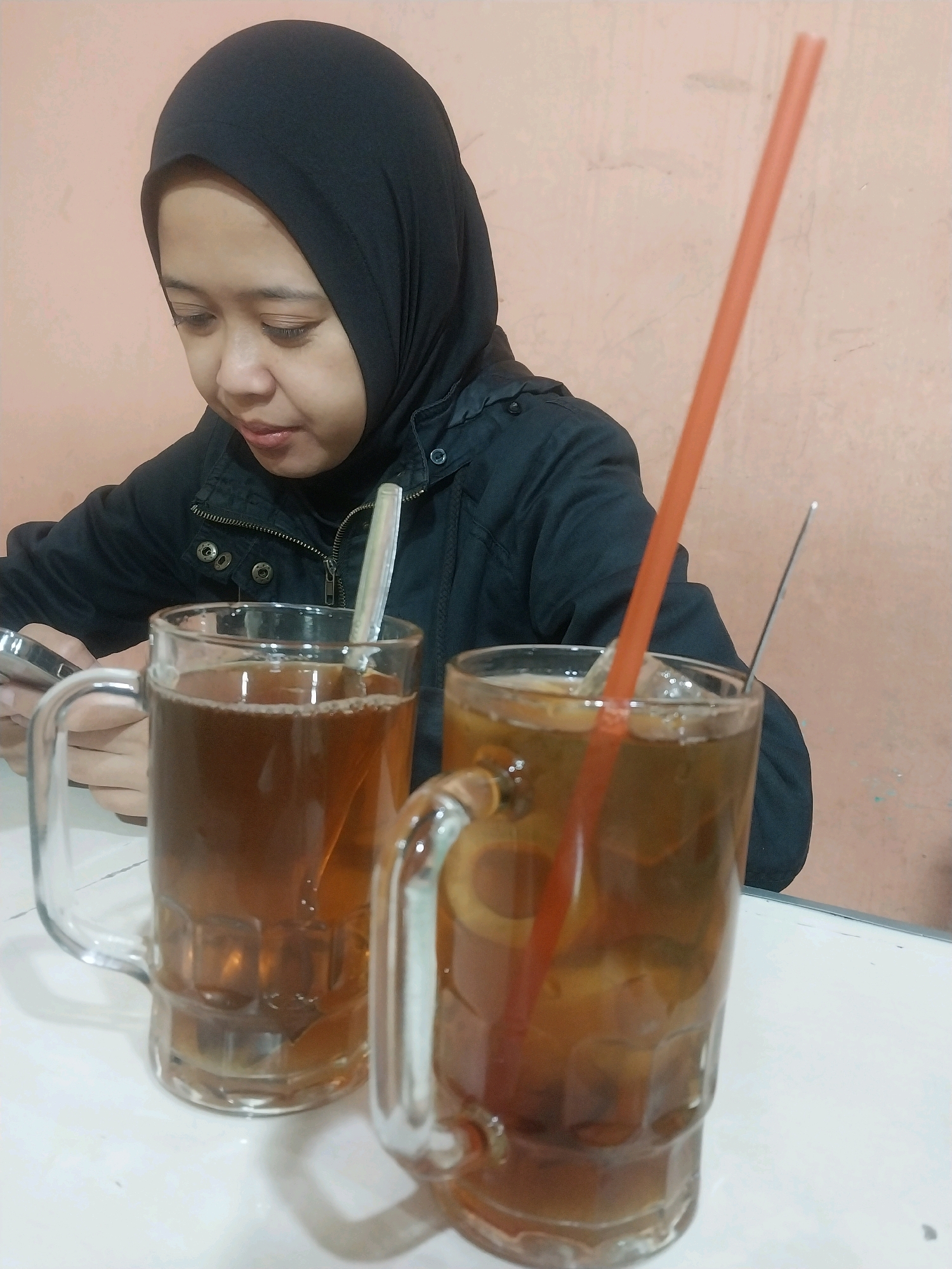 In Sleman, Indonesia, a young woman wearing a black hijab and a black jacket is seated at a table, looking down and to her left, possibly at a phone. Two glasses of iced tea are in front of her. The glass on the left contains tea and a spoon, while the glass on the right contains tea, ice, and a red straw. The table surface is white, and the background wall is a light peach color.