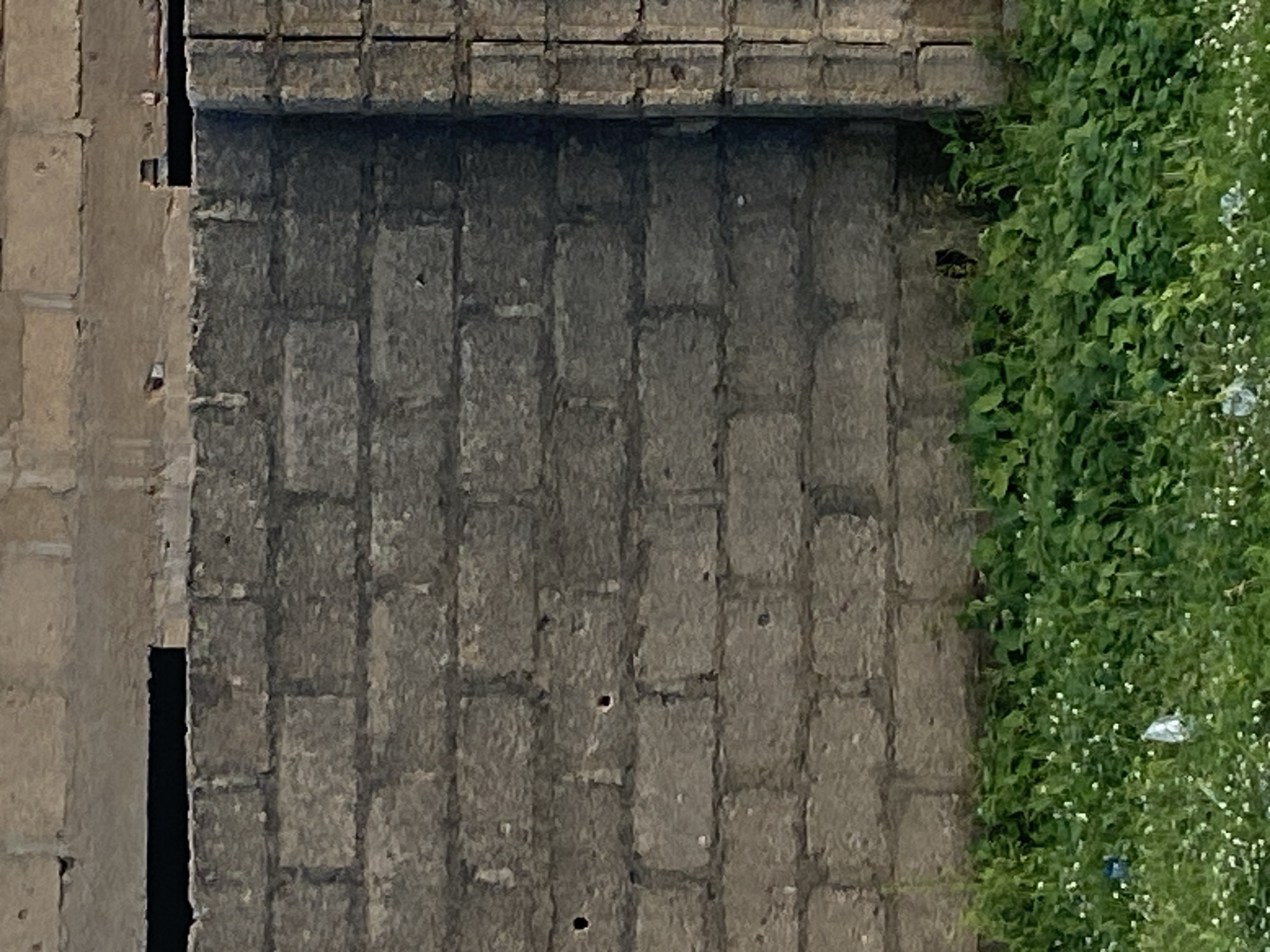 A low-angle, full-shot photograph shows a thick, grey cement block wall that runs horizontally across the frame. The wall is constructed of interlocking blocks and has several small, dark holes scattered across its surface. The top of the wall is partially obscured by a lighter-colored structure made of larger, more irregular blocks. A horizontal dark rectangular opening is visible near the top of the frame, cutting across the lighter-colored structure. To the right of the main wall, a narrower, vertical section of similar grey blocks is visible. In the foreground, a dense patch of lush green vegetation with small white flowers grows at the base of the wall. Some white plastic litter is visible within the greenery. The scene is set in Jauro Yinu, Nigeria.