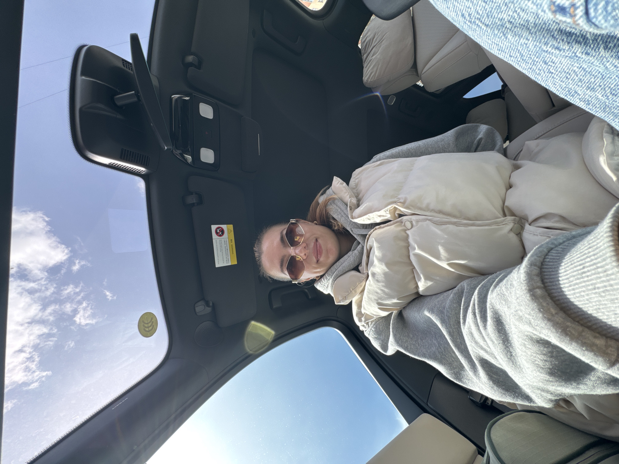 A low-angle interior shot shows a woman wearing sunglasses and a white puffer vest over a gray hoodie. She is seated inside a car, likely in the passenger seat, as indicated by the overhead console and a sun visor with safety information. The car's sunroof provides a view of a blue sky with scattered clouds. A yellow sticker is visible on the inside of the windshield. Portions of the car's seats and interior are visible. The location is unknown.