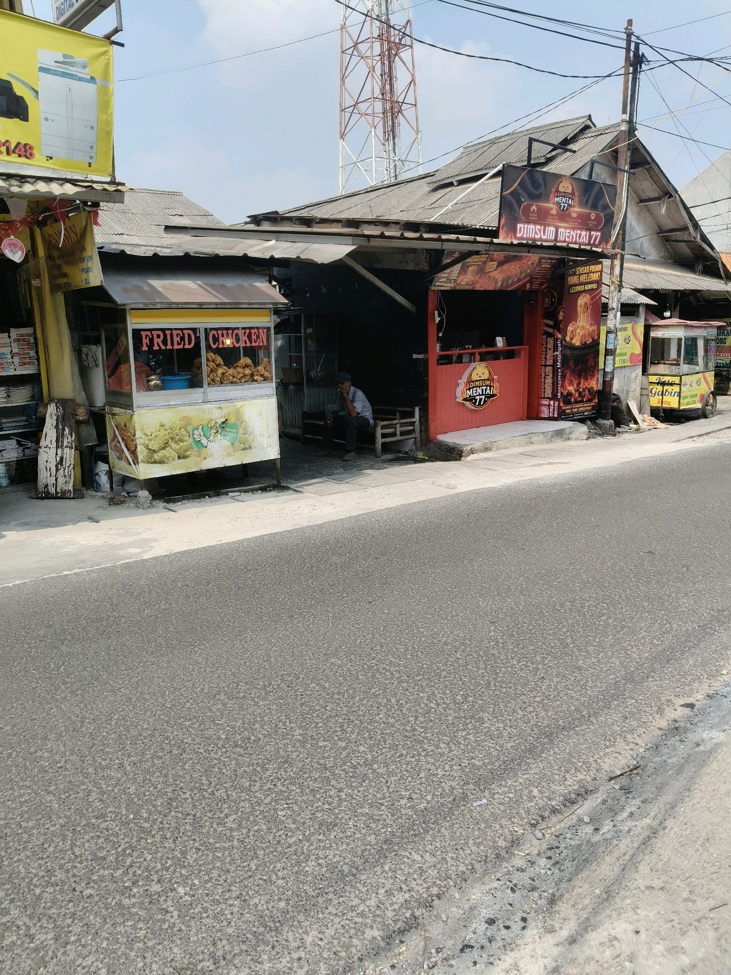 The image captures a street scene in South Tangerang, Indonesia, featuring several small food stalls. On the left, a stall with a yellow banner displays a large image of a photocopier and the text "2148". Adjacent to it is a "Fried Chicken" stall with a glass display case filled with fried chicken and signage that includes a cartoon chicken. Further down the street, a stall with a red facade displays a "Dimsum Mentai 77" sign. Another stall, identified as "Kue Gabin", is visible on the right, with a wheeled cart. A man is seated on a bench outside the "Dimsum Mentai 77" stall. The stalls are situated along an asphalt road under a bright, clear sky. A telecommunications tower is prominent in the background.
