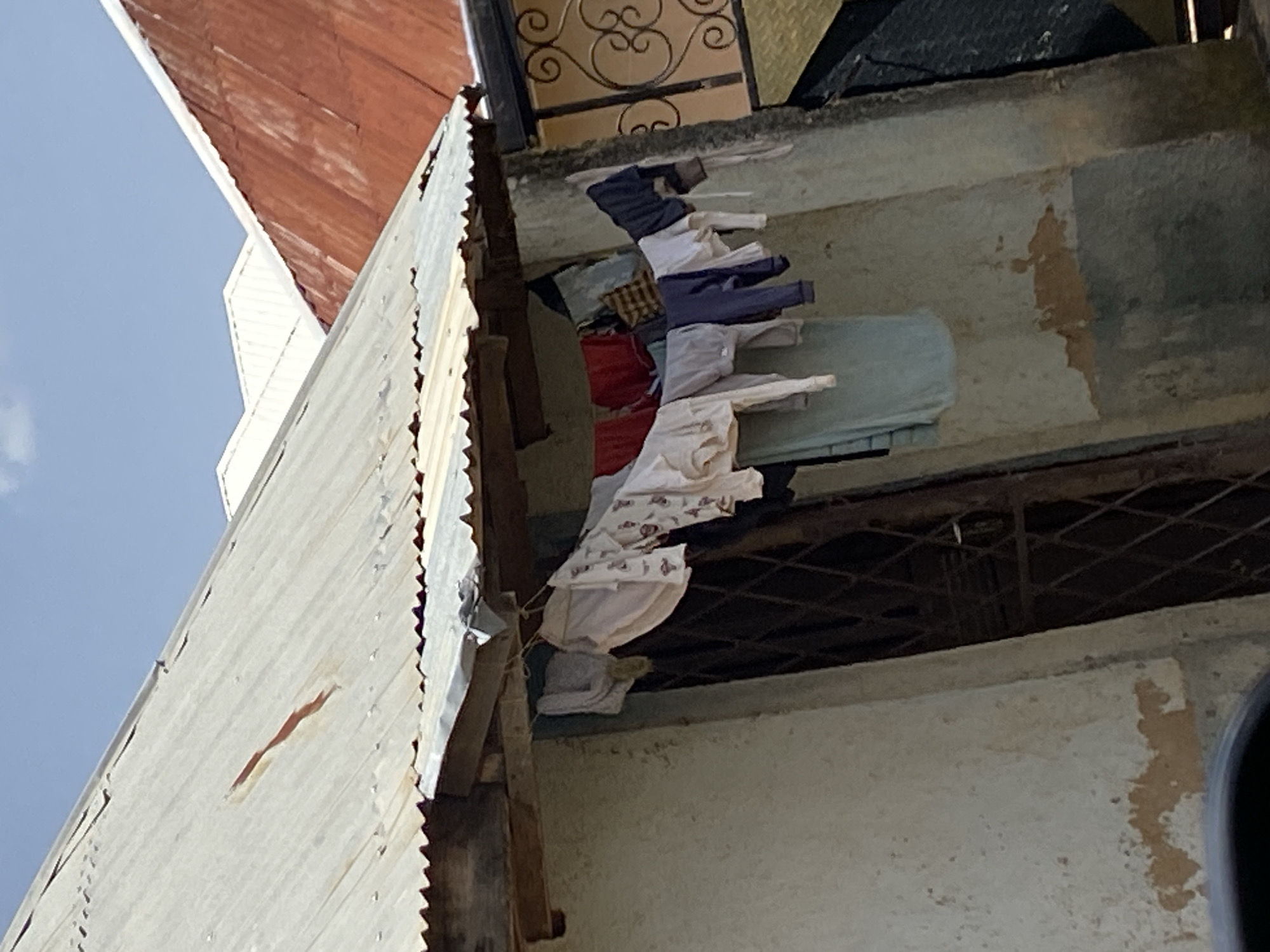 The image depicts an outdoor scene in Bar Jirgi Summa, Nigeria. In the foreground, clothing hangs on a clothesline attached to the underside of a corrugated metal roof. The clothing includes various items such as shirts and what appear to be trousers. The building beneath the roof has textured, weathered walls, and a barred doorway is visible. In the background, several rooftops are visible, including a red corrugated metal roof and sections of white roofing. The sky above is a clear blue. The overall impression is of a residential or domestic setting in a somewhat rural or less developed urban area.