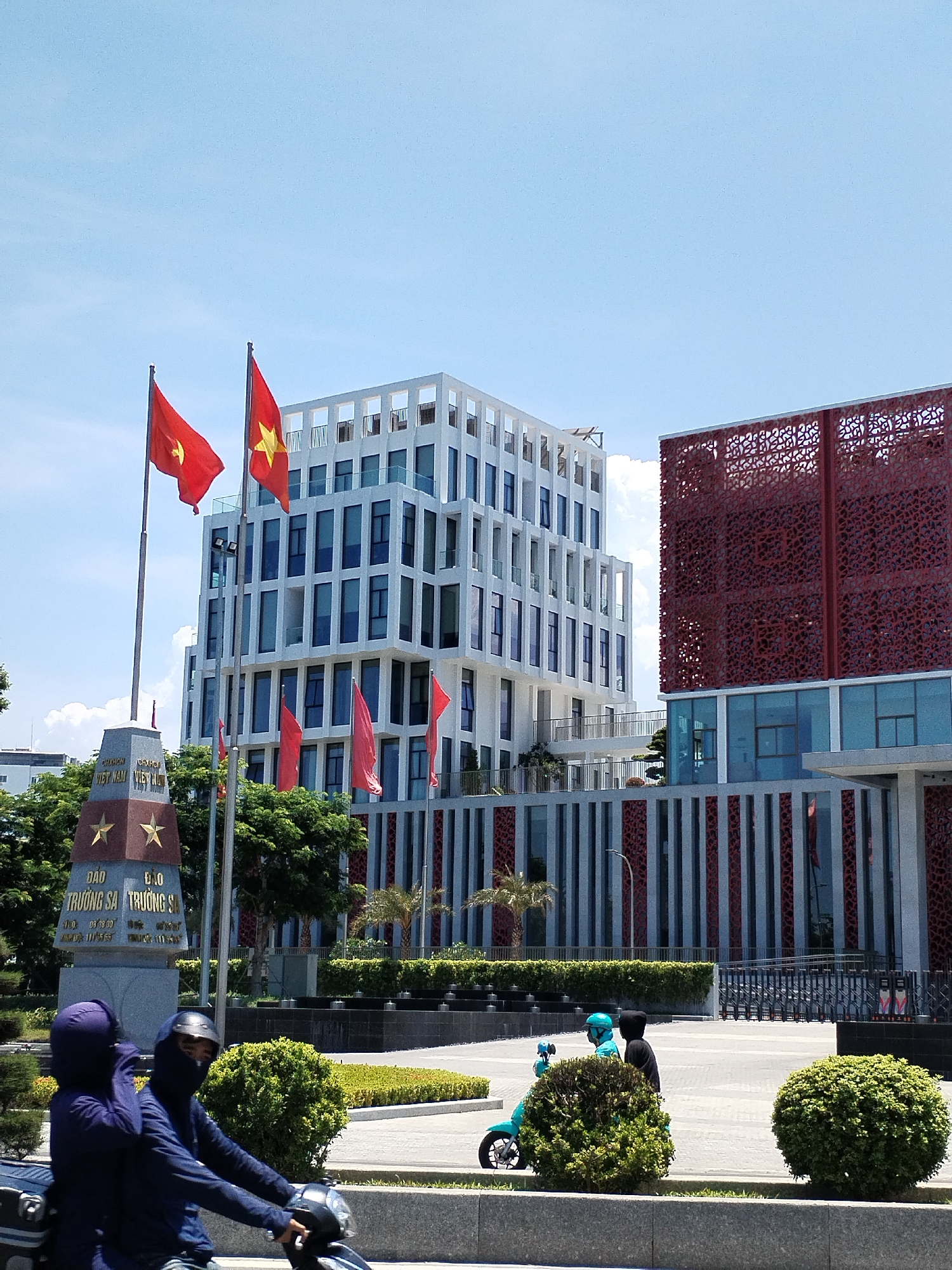 In Nha Trang, Vietnam, on a sunny day with a bright blue sky and scattered clouds, a prominent monument stands on the left, topped with Vietnamese flags. Behind it, a modern, multi-story white building with a lattice-like exterior and numerous windows dominates the scene. To the right of this building, another structure features a striking red, patterned facade. In the foreground, two individuals on a light blue scooter are visible, their faces partially obscured. Further to the left, two people on a motorcycle, one wearing a dark blue outfit and the other a dark jacket and helmet, are also present. The area is landscaped with green bushes and trees, suggesting a public space or plaza.