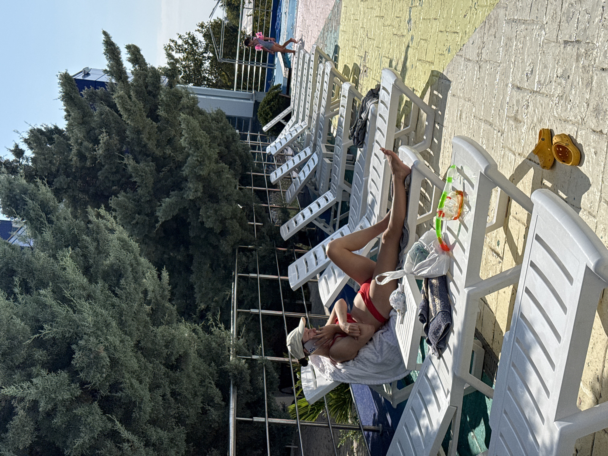 A woman in a red bikini and a white cap is relaxing on a white lounge chair by a swimming pool. She is holding a phone and looking at it. Beside her on the chair is a towel and a pair of goggles. There is a line of empty lounge chairs behind her. In the background, a child in a pink swimsuit and life vest is walking on a platform. The scene appears to be a sunny day, possibly in the afternoon given the long shadows cast by the lounge chairs. The setting is an outdoor pool area with a blue tiled pool and a yellow-tiled deck. There are also large evergreen trees visible to the left. The overall mood is one of leisure and relaxation. The image is a vertical shot, with the subjects oriented as if the camera is tilted.
