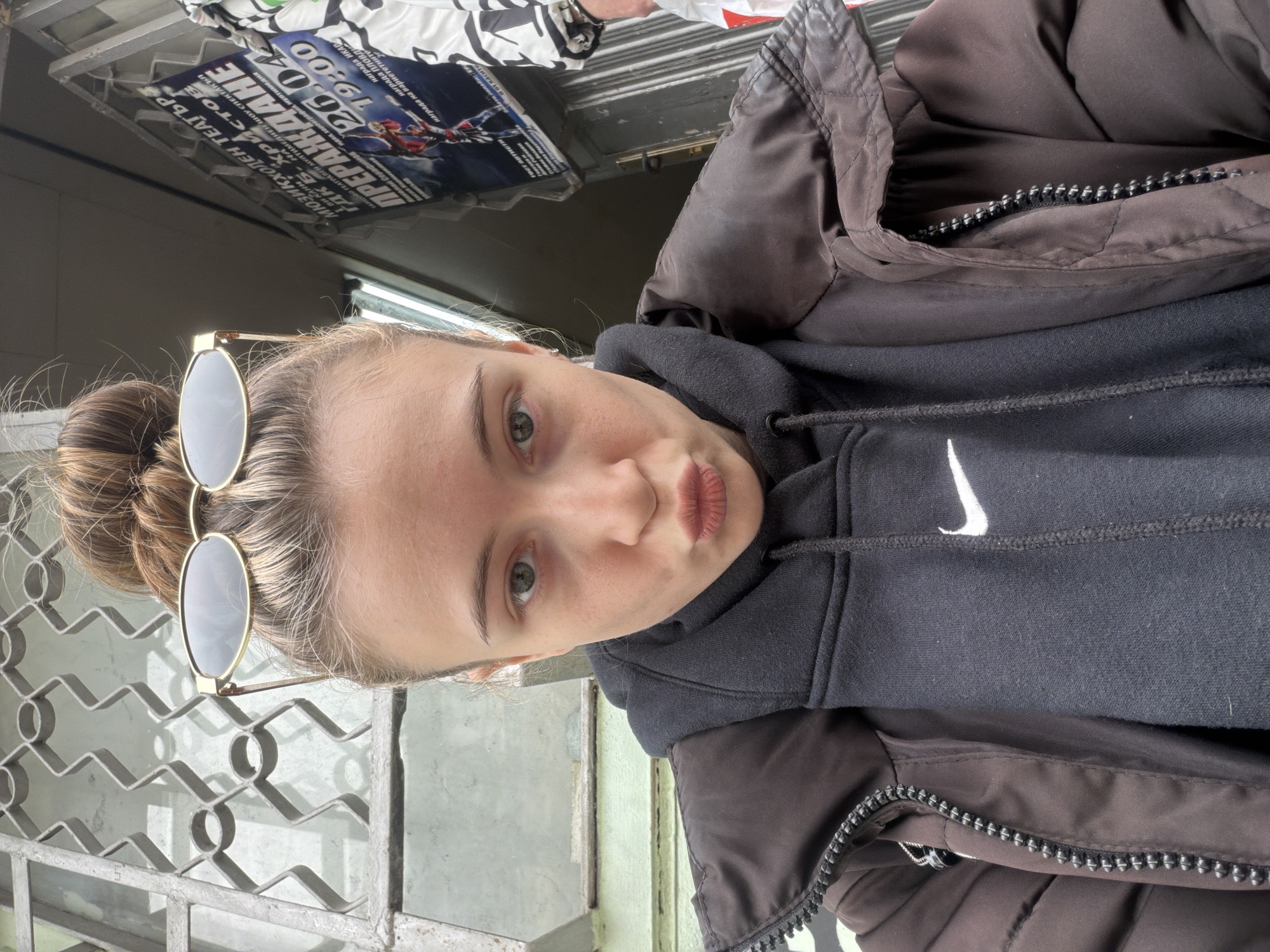 A young woman with light skin and blonde hair styled in a bun is posing for a selfie outdoors. She is wearing a dark brown puffy jacket over a black Nike hoodie. Sunglasses are perched on her head.
The background includes a metal fence, a light-colored wall, and a poster. The poster mentions "Мюзикхолен Театър", is associated with the city of Plovdiv, Bulgaria and refers to a show or event titled "Прераждане" scheduled for April 26th at 19:00.