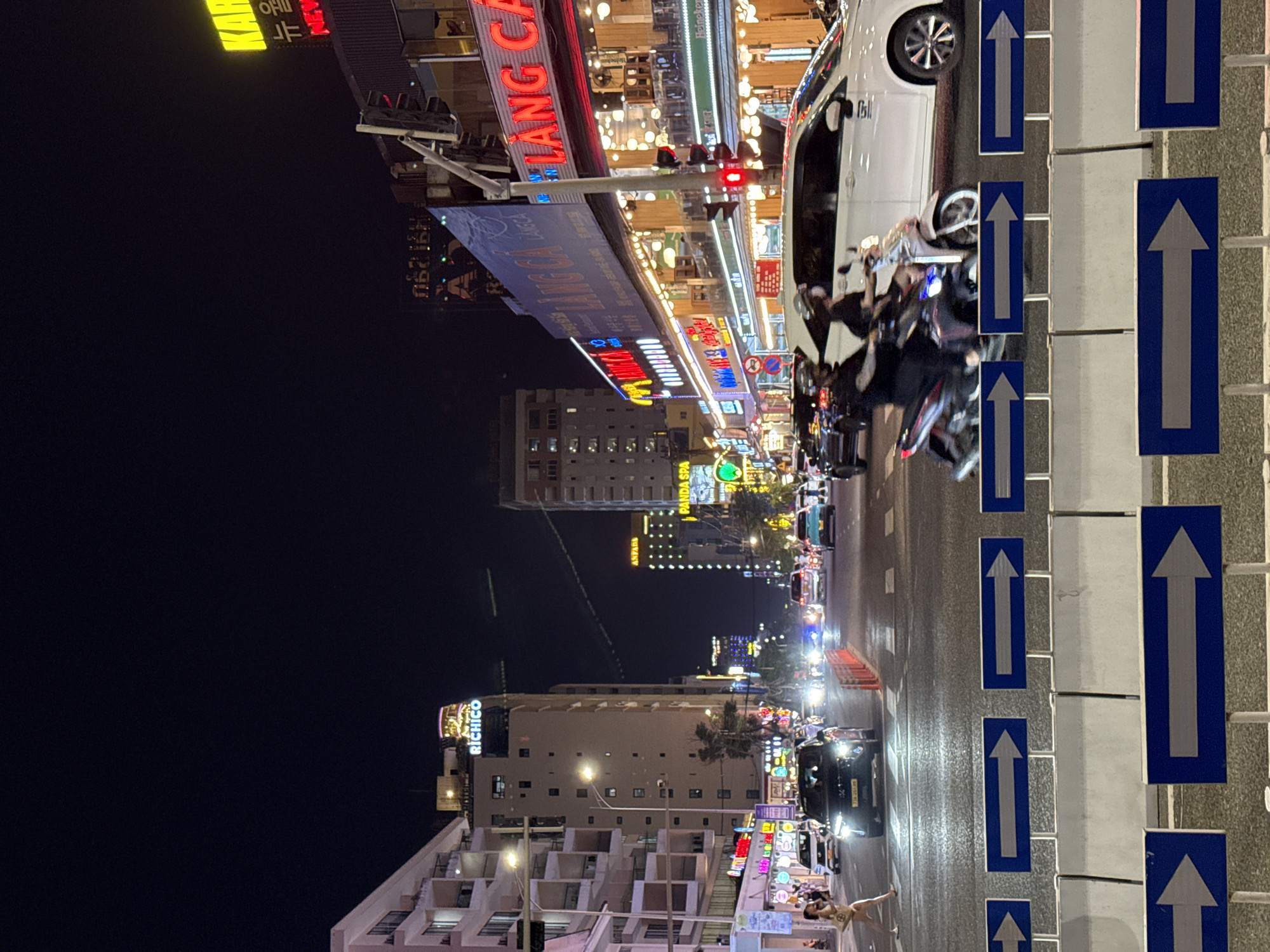 The image depicts a bustling street scene at night in Đà Nẵng, Vietnam. The perspective is tilted, with the street and buildings appearing to run vertically. The scene is illuminated by a vibrant array of neon signs and streetlights, indicating it is nighttime.
The street is filled with traffic, primarily motorcycles and a few cars. The motorcycles are packed closely together, suggesting a busy urban environment. On the right side of the street, a row of blue and white directional signs with white arrows pointing upwards are prominently displayed, guiding traffic. A white car is parked on the left side of the street, and a group of people on motorcycles are maneuvering past it.
The buildings lining the street are commercial establishments, with various businesses indicated by illuminated signs. One sign clearly reads "RICHICO" in large, glowing letters. Other signs display a mix of text and images, suggesting shops, restaurants, or entertainment venues. The architecture of the buildings appears to be modern, with multiple stories.
In the foreground, towards the bottom left, a person is visible standing on the sidewalk, seemingly posing for the photo. Their attire is light-colored and casual. The overall atmosphere of the scene is energetic and lively, typical of a vibrant city at night. The wet appearance of the road surface suggests recent rain or that the street has been recently washed.