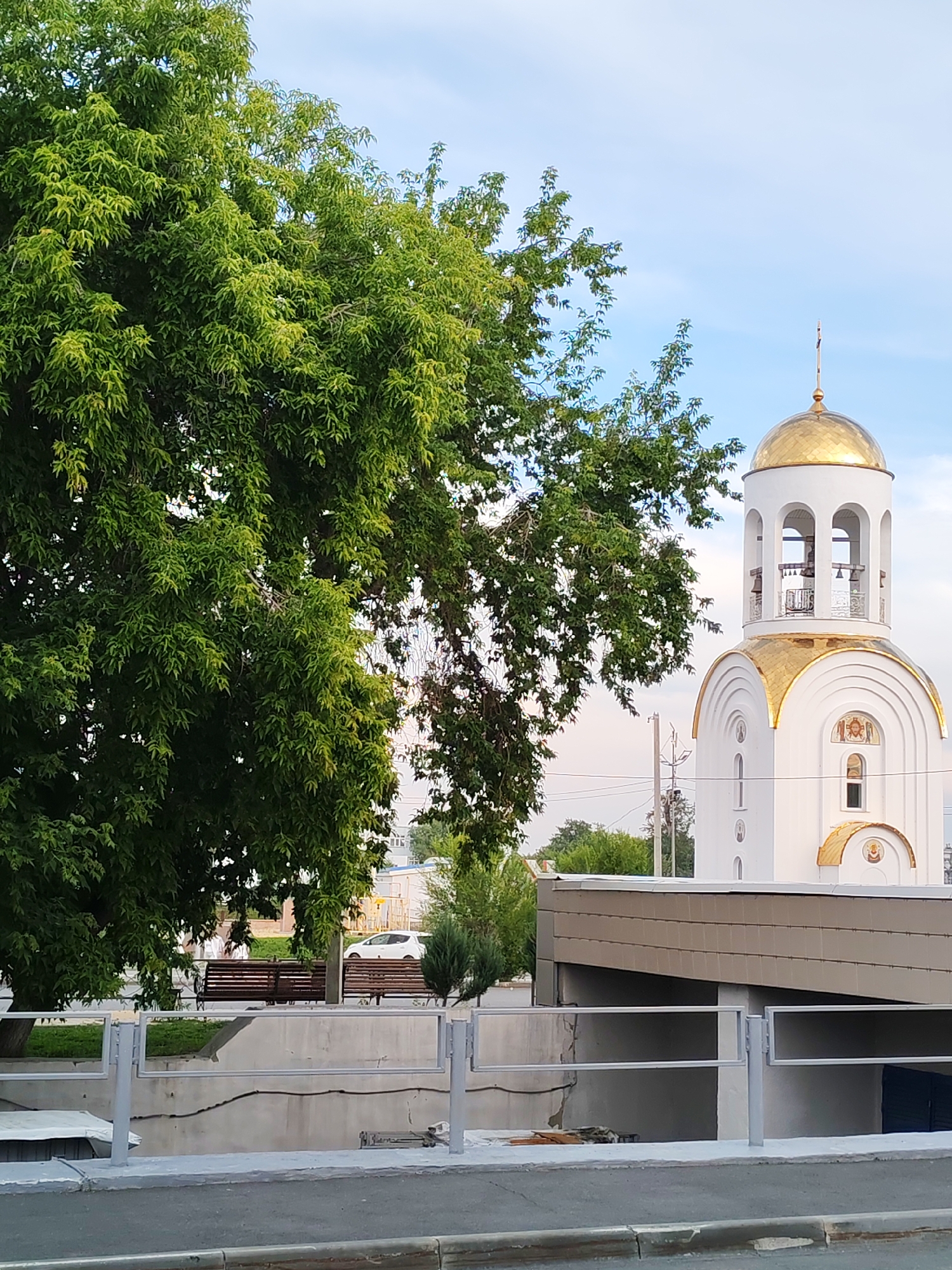 A white church with a gold dome and cross stands on the right side of the image, partially obscured by a large green tree on the left. The church has arched windows and decorative religious imagery. In the background, there are other trees, power lines, and a white car. In the foreground, there is a modern building with a textured facade, a metal fence, and a paved area that appears to be a street or sidewalk. Two wooden benches are visible on a grassy area behind the fence. The sky is partly cloudy, suggesting it is daytime. The scene appears to be a public space in Kurgan, Russia. There is no visible text.