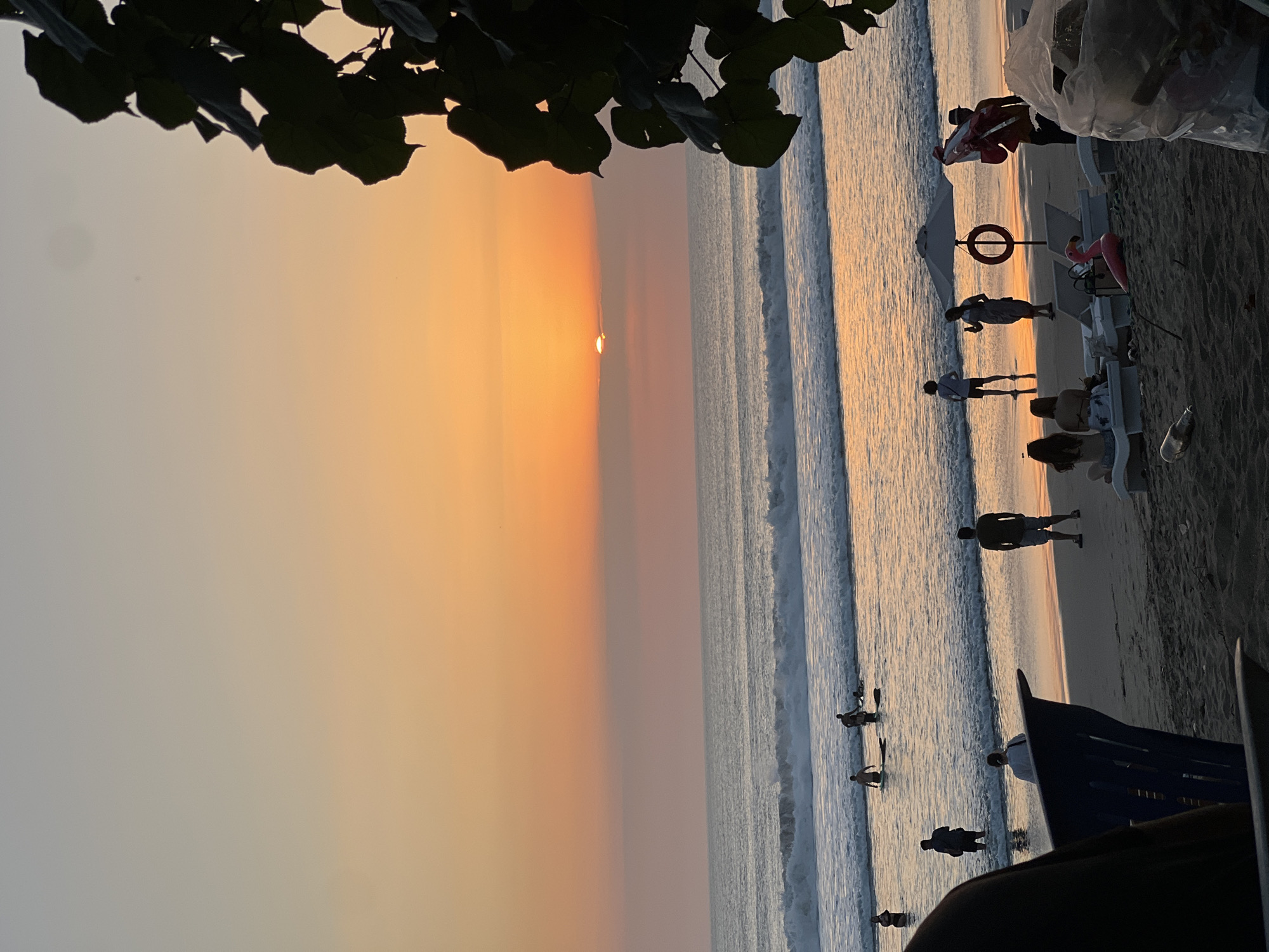This image depicts a sunset scene on Kuta Beach, Bali, Indonesia. The sky transitions from a pale blue at the top to a vibrant orange and pink near the horizon where the sun is partially obscured by clouds. The ocean displays gentle waves with white foam, reflecting the warm hues of the sunset.
On the beach, several individuals are present. In the water, a group of people are seen, some with surfboards. Closer to the shore, other people are standing or walking in the shallows. On the sand, further back from the water, there are more people. Some are seated on beach chairs, while others are standing. Notable objects include beach chairs, a large white umbrella, a lifebuoy hanging from the umbrella, and a pink inflatable flamingo. In the foreground on the right, a large plastic bag filled with trash is visible, along with a plastic bottle on the sand. On the left, a portion of a dark-colored chair is in the foreground. Foliage from a tree in silhouette frames the upper right portion of the image.