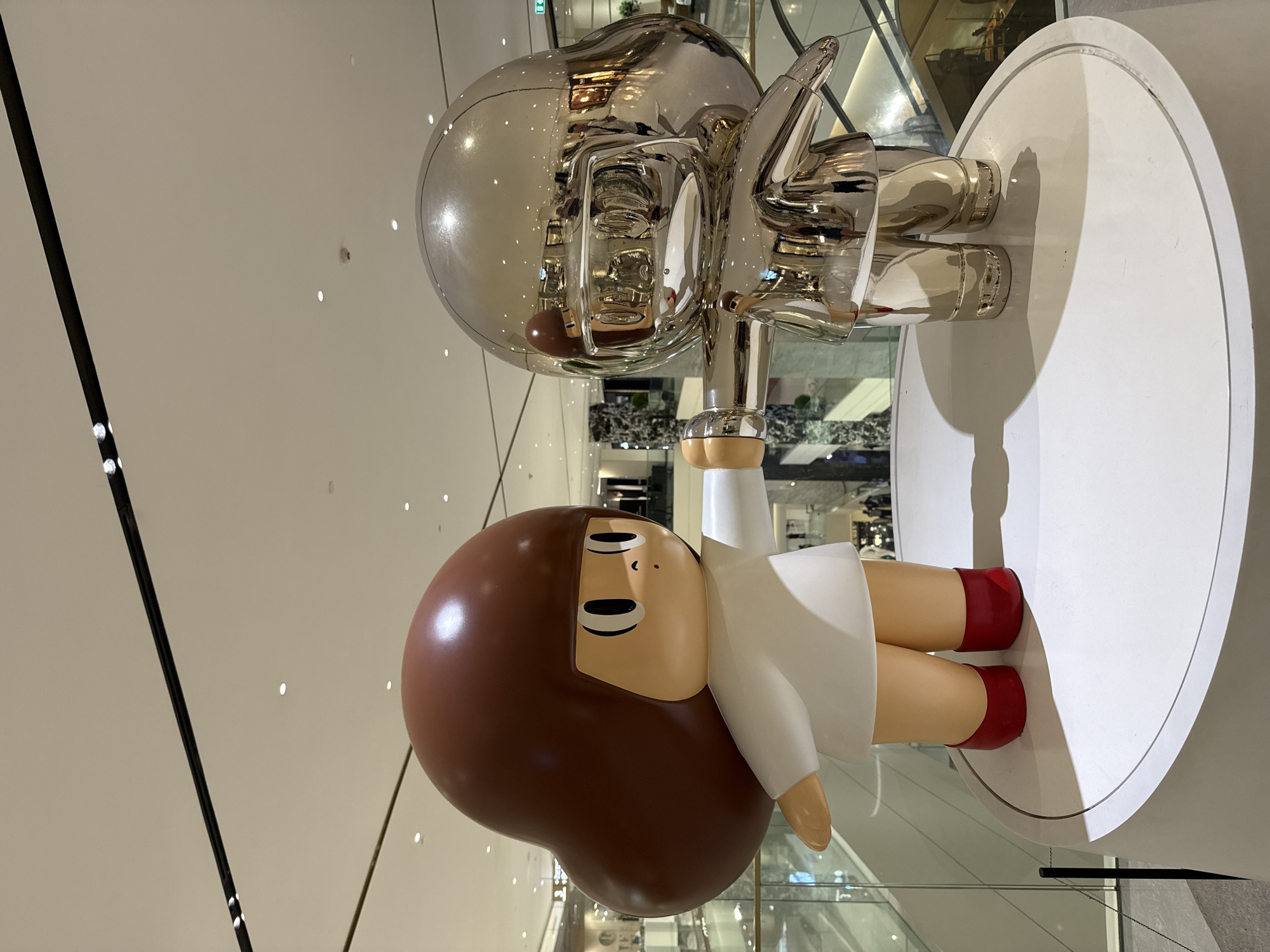 A low-angle photograph depicts two adjacent modern art sculptures inside a building in Bangkok, Thailand. The sculpture in the front is a glossy, brown-haired girl wearing a white dress and red shoes. She is lying on her stomach with her arm raised. Behind her is a larger sculpture of a silver, reflective figure giving a thumbs-up. Both sculptures are mounted on a white, circular pedestal. The surrounding environment appears to be a shopping mall with polished floors and high ceilings. Reflections of the mall’s interior are visible on the surface of the silver sculpture.